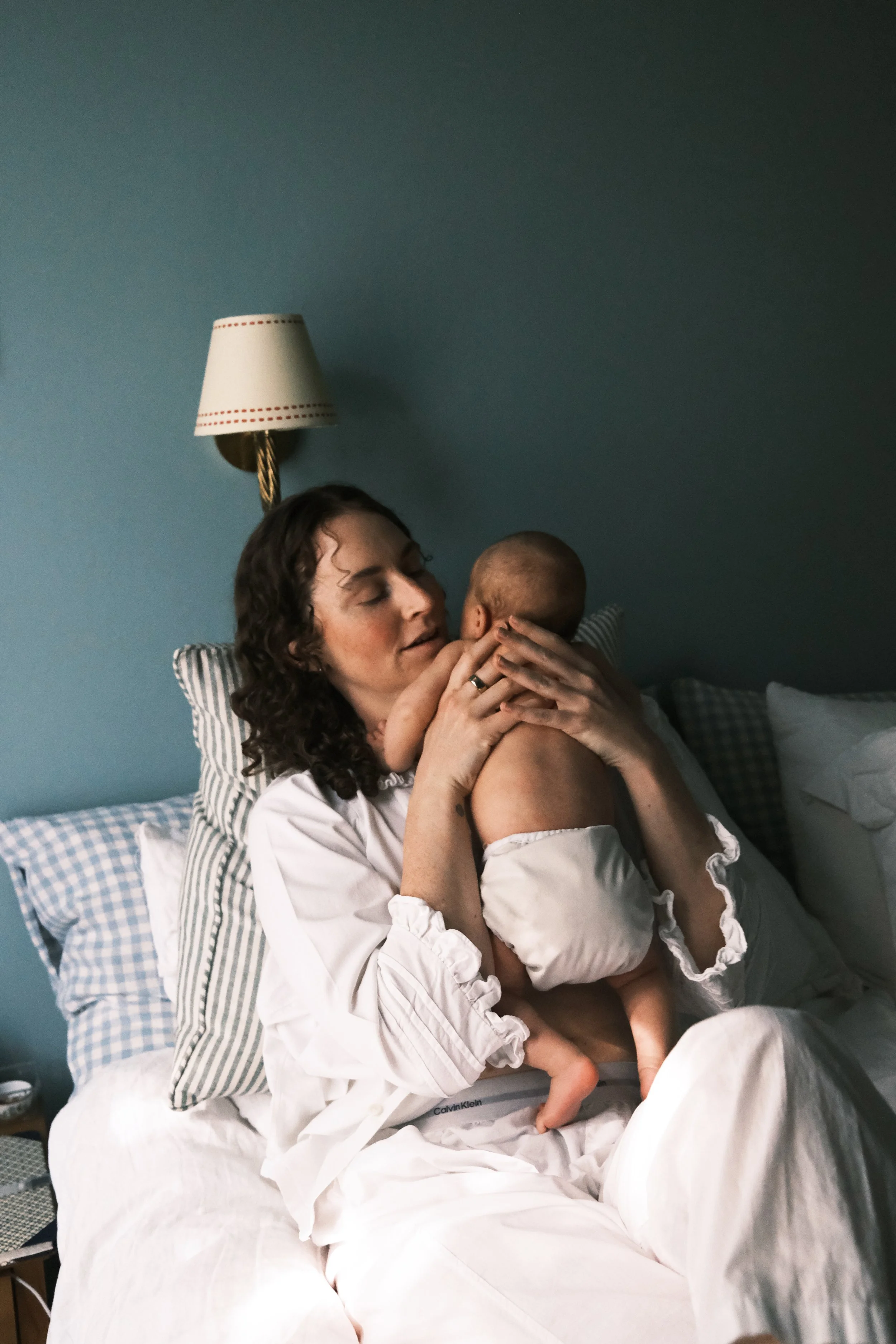 A woman with curly brown hair, wearing a white button-up shirt, sitting on a bed, holding and kissing a small baby. The bed has striped pillows and a blue wall with a hanging lamp in the background.