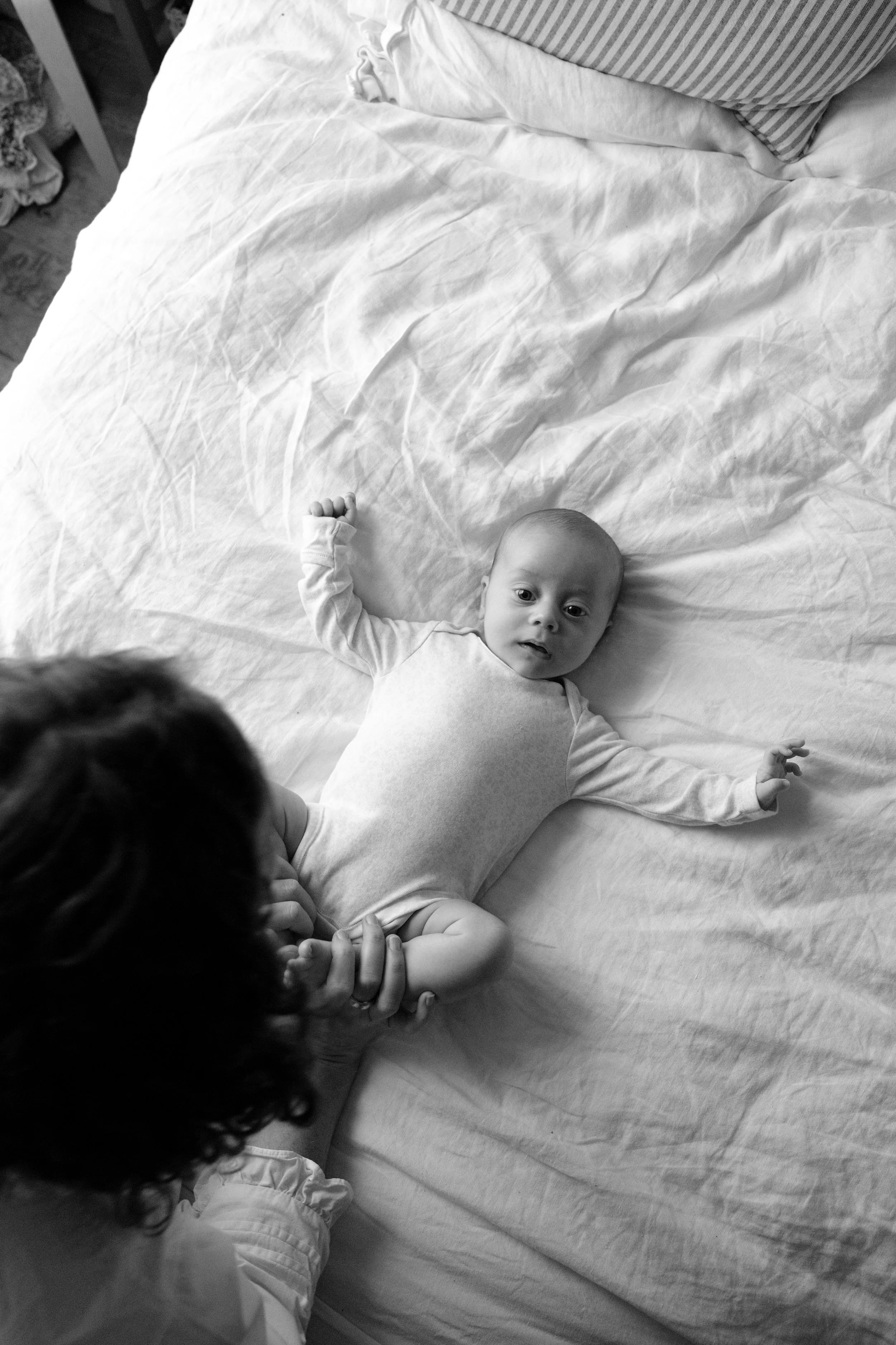 A baby lying on a bed while an adult holds the baby's hand. The baby is looking towards the camera, and there is a pillow with striped pillowcase in the background. The photo is in black and white.