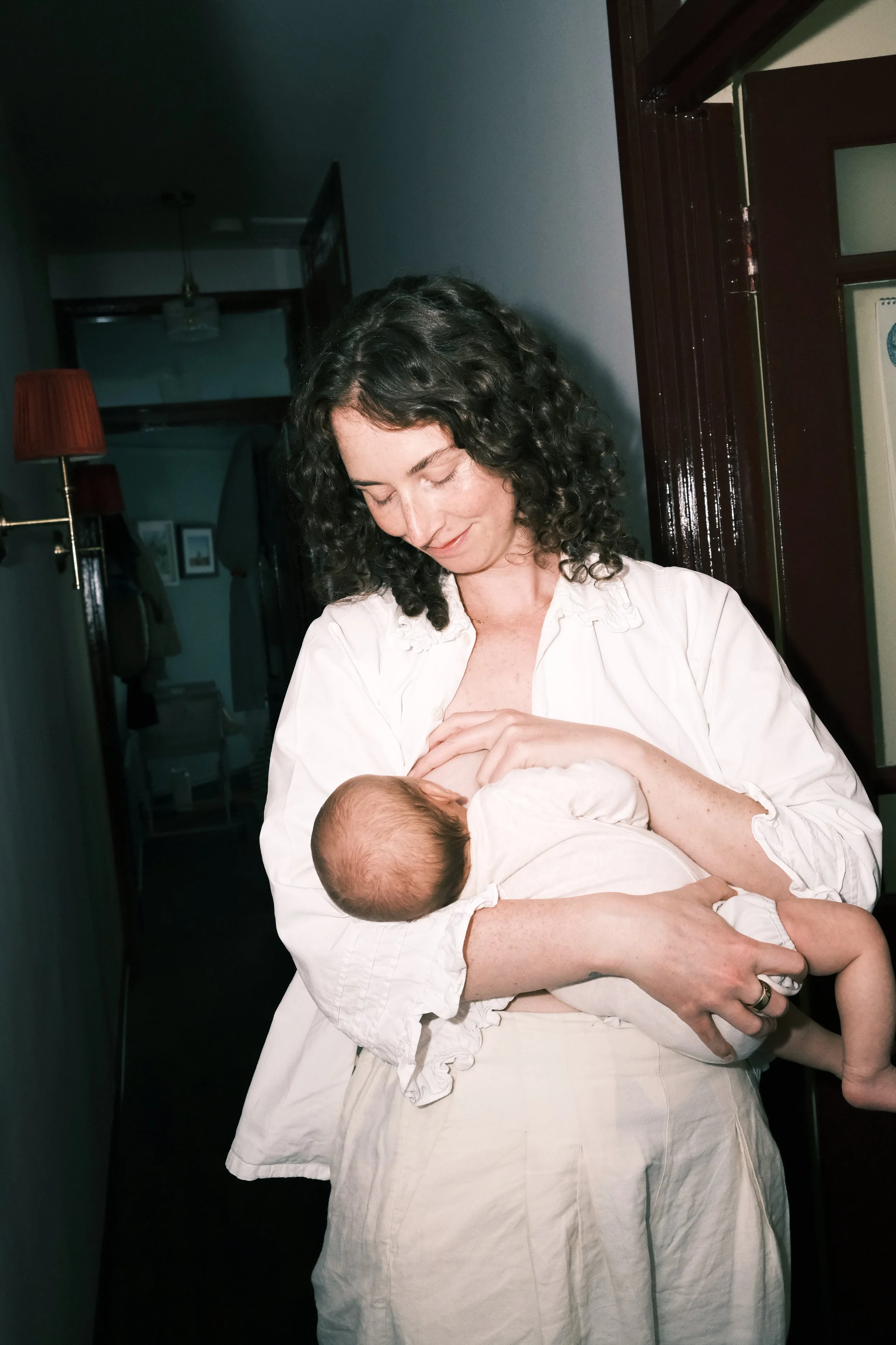 A woman with curly dark hair is smiling gently while holding a baby close to her chest. The woman is wearing a light-colored blouse and a white skirt, and the baby is dressed in light-colored clothing. They are standing indoors near a doorway, with a