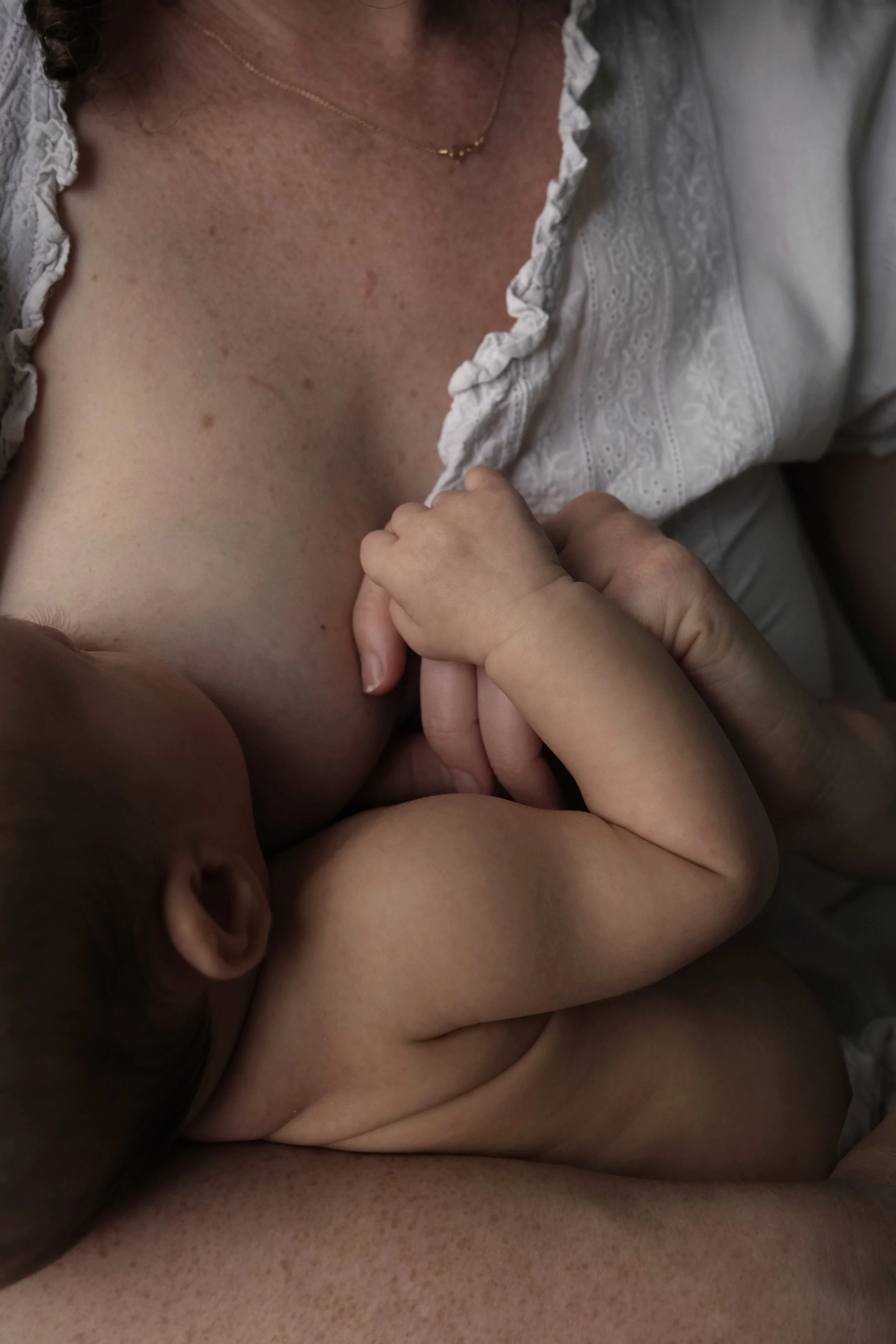 Close-up shot of a woman in a white embroidered blouse, wearing a gold necklace, and holding a child's hand, with part of the child visible and lying on the woman's chest.