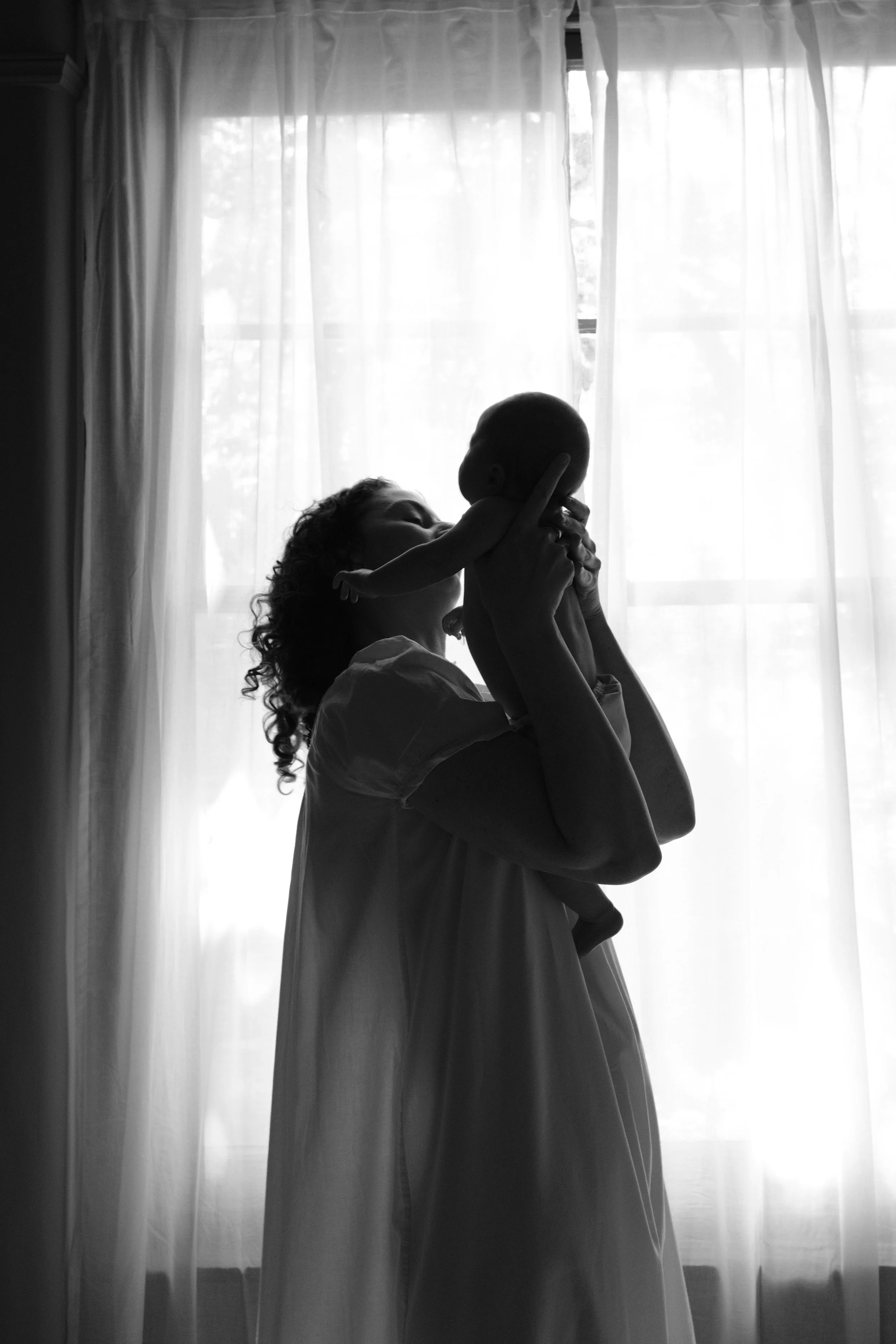 A person holding a baby in front of a sheer curtain with sunlight streaming in.