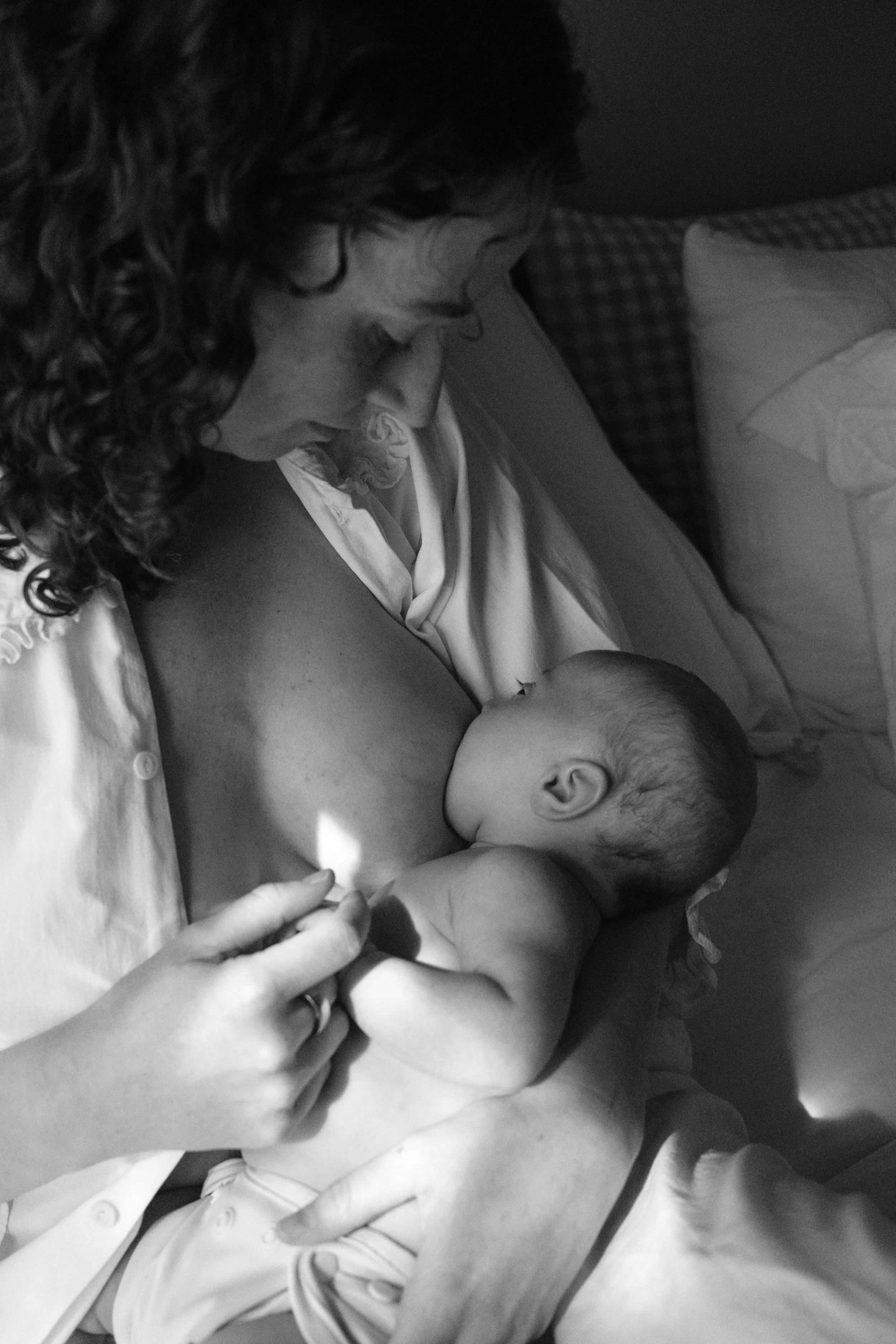 A woman with curly hair breastfeeding a baby in a hospital bed, holding the baby's head and supporting their body with her arm.
