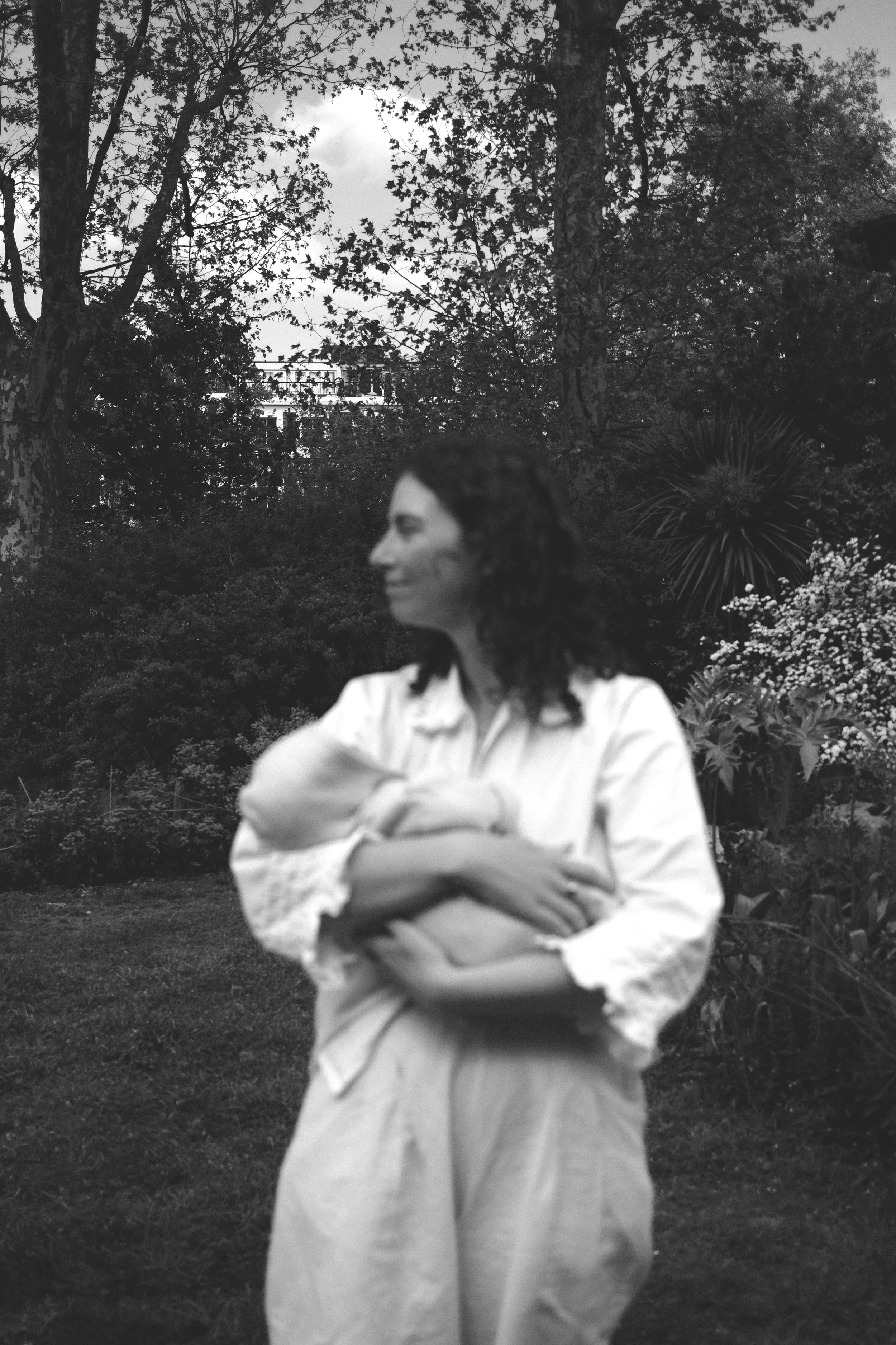 A woman with curly hair standing outdoors in a garden or park, with trees and plants in the background, wearing a white blouse and pants, arms crossed, looking to the side.