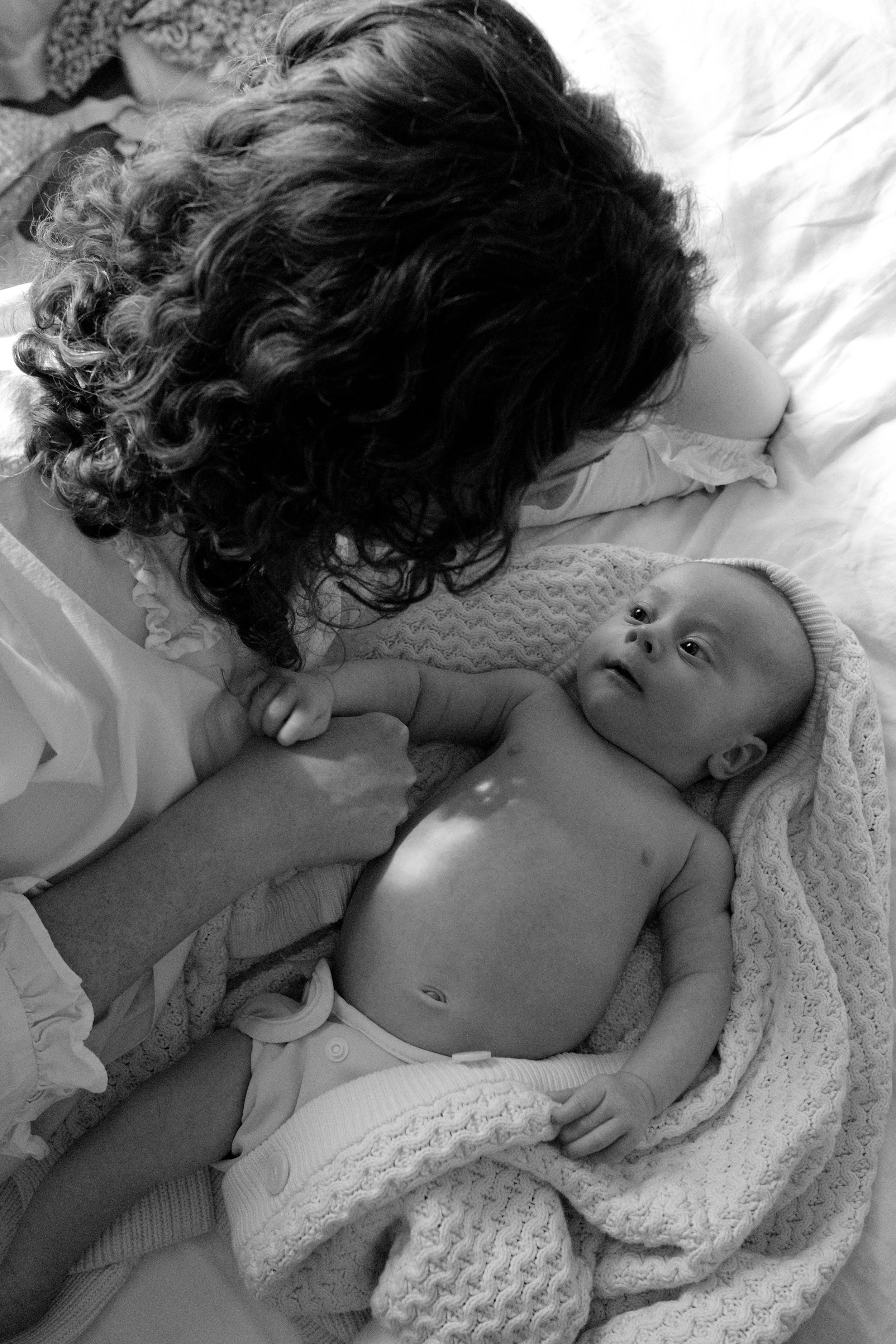 A newborn baby is lying on a bed wrapped in a knitted blanket, looking up with wide eyes, while a woman with curly hair is lying next to the baby, holding the baby's hand and looking at the infant.