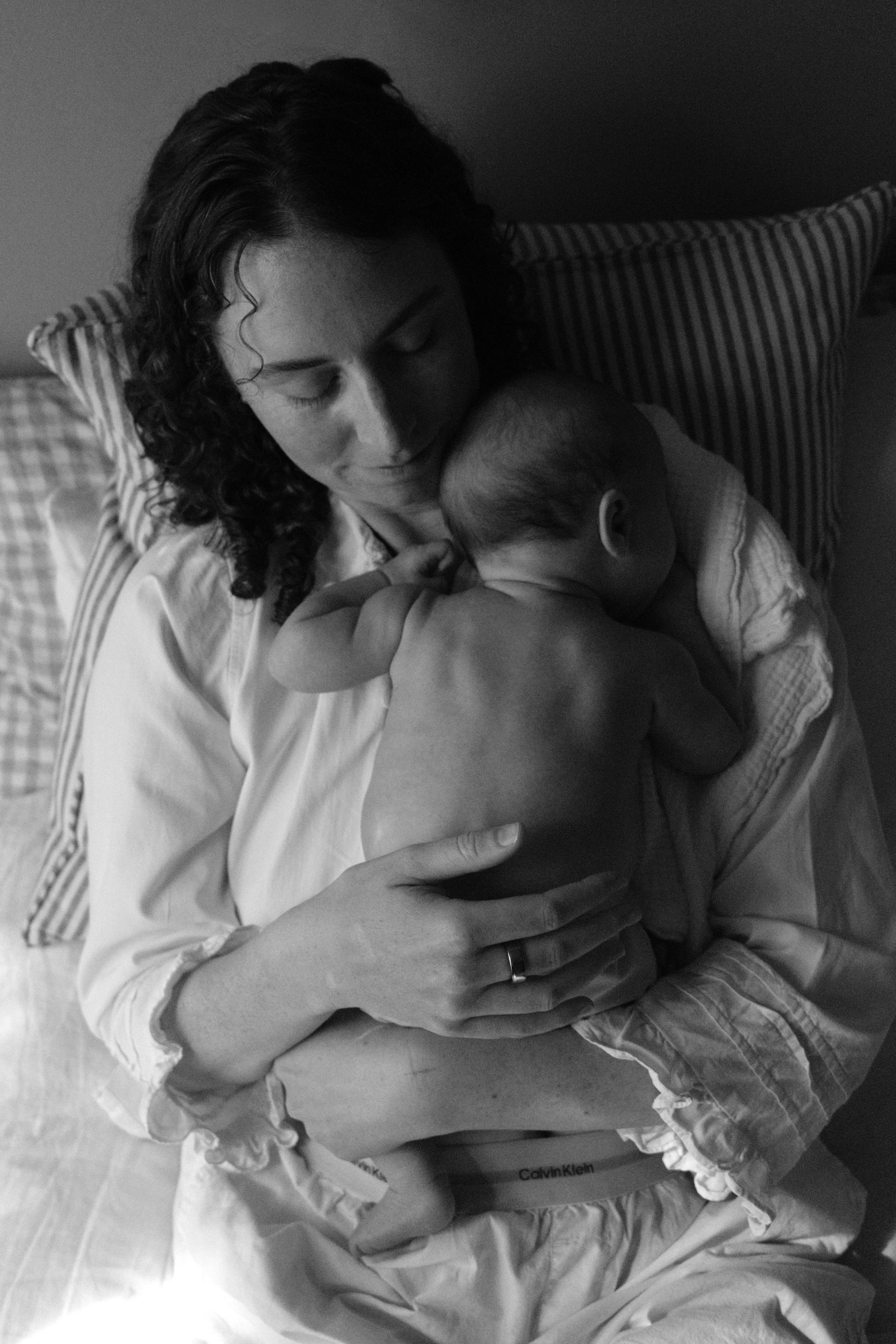 A woman with curly hair feeding and cuddling a baby in a crib or bed, with the woman smiling softly and the baby resting peacefully.