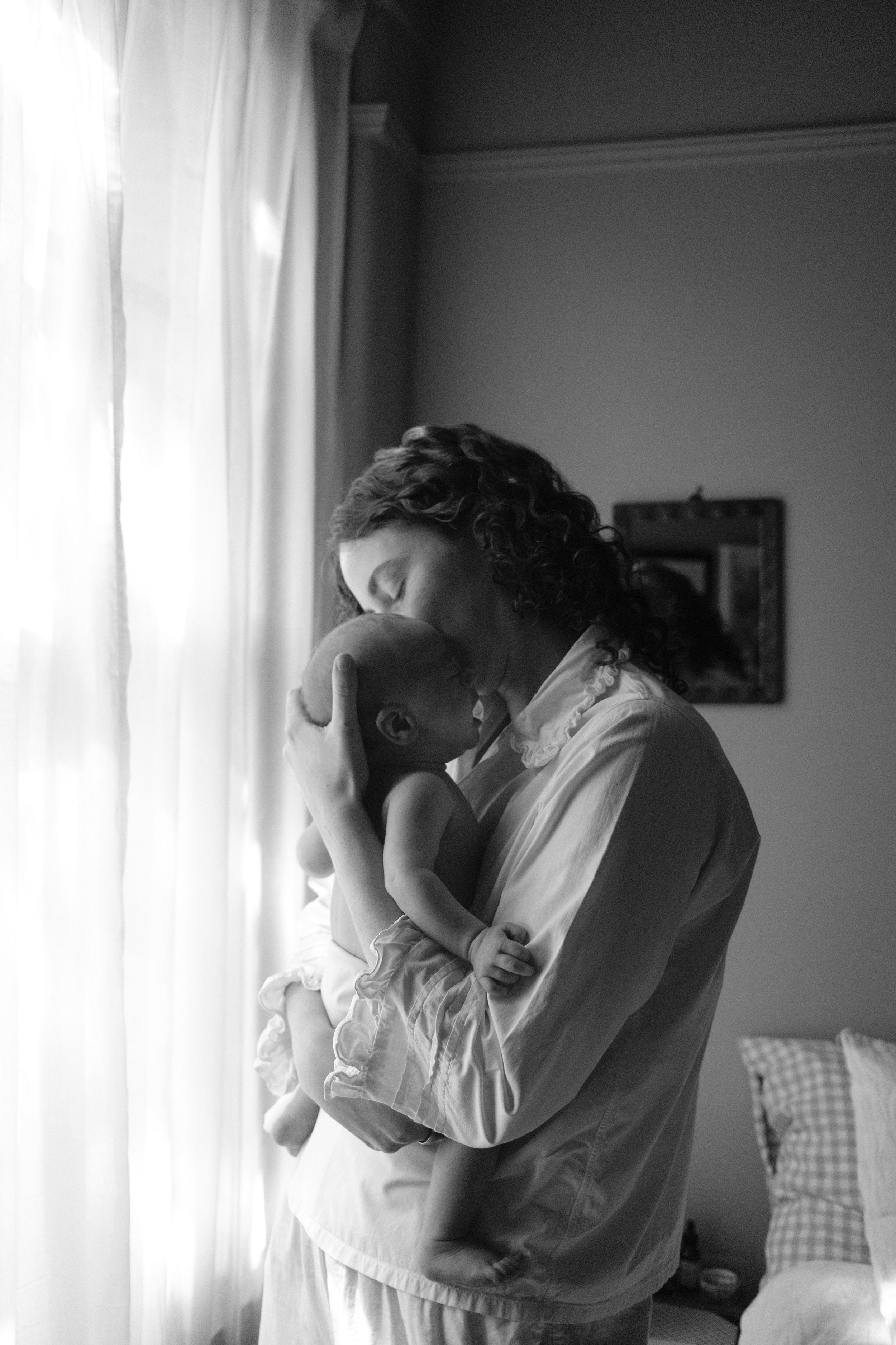 A woman kisses a baby on the forehead while holding the child in front of a window with light curtains, in a bedroom with a bed and pillows in the background.