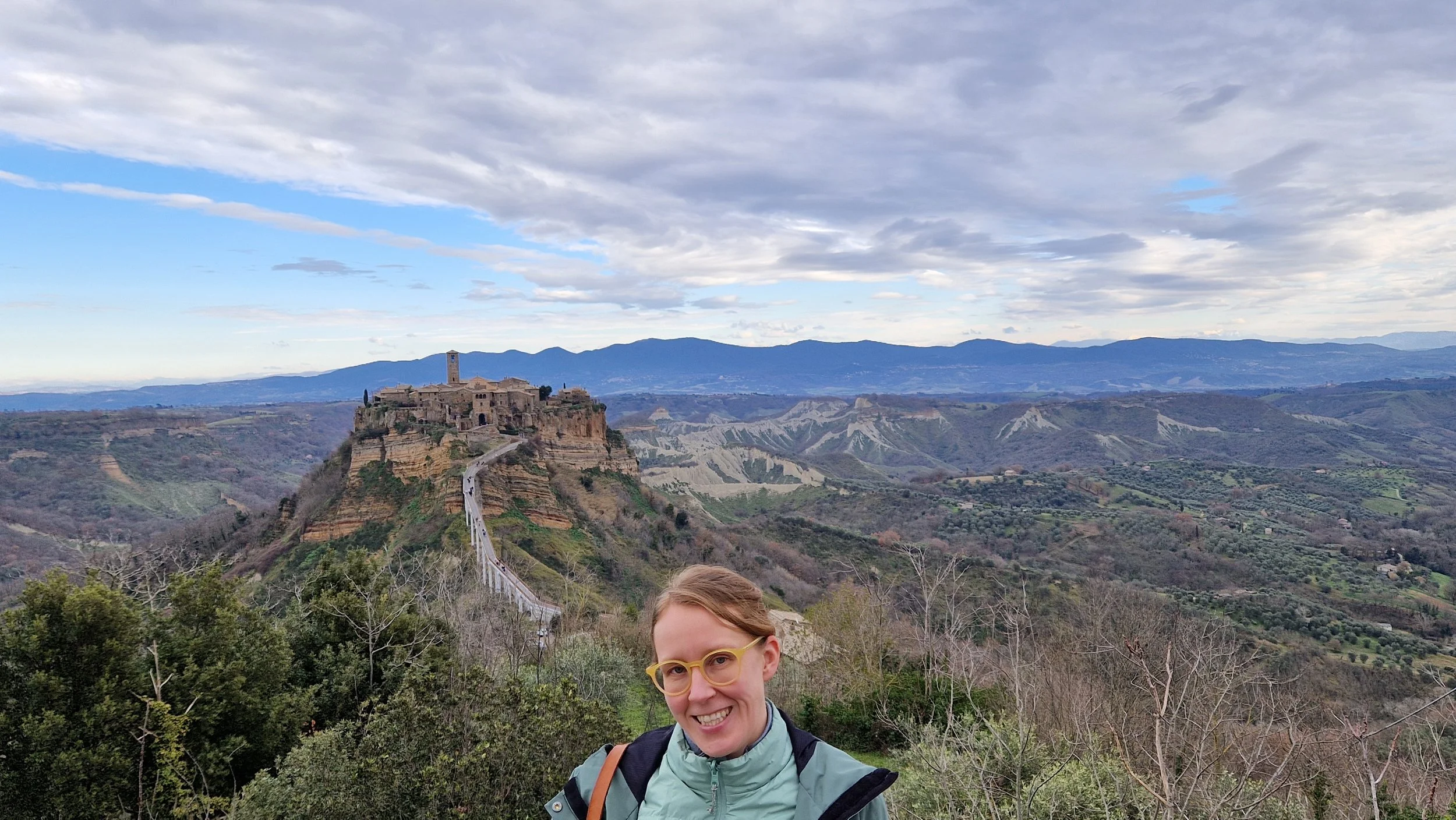 Kuvan edessä hymyilevä nainen (Eeva-Sofia Anttonen) ja taustalla näkyy kapea laaksossa kulkeva tie, joka vie pieneen kylään, joka on rakennettu kallion päälle.