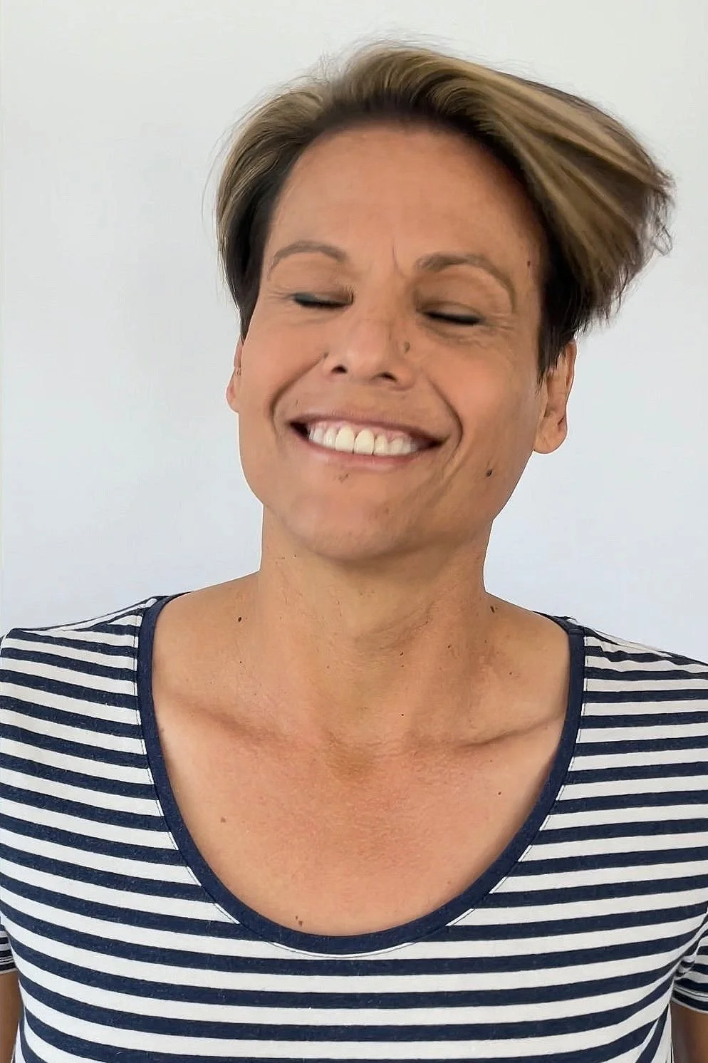 A woman with short, light brown hair smiling with her eyes closed, wearing a navy blue and white striped shirt, standing against a plain white background.