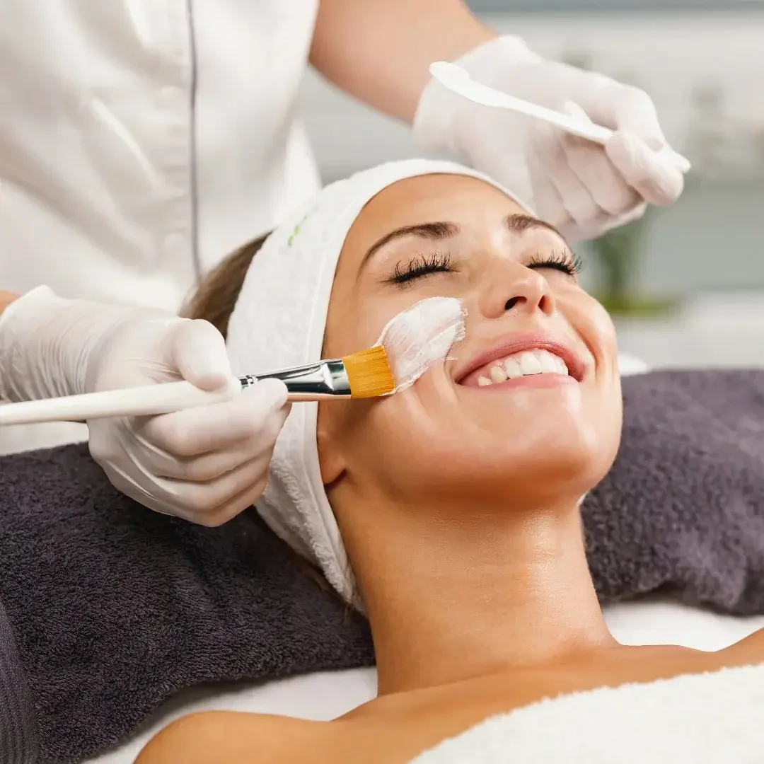 A woman is receiving a facial treatment at a spa or skincare clinic. She is lying down with a towel on her head and a smile on her face. A skincare professional is applying a white facial mask or cream with a brush.