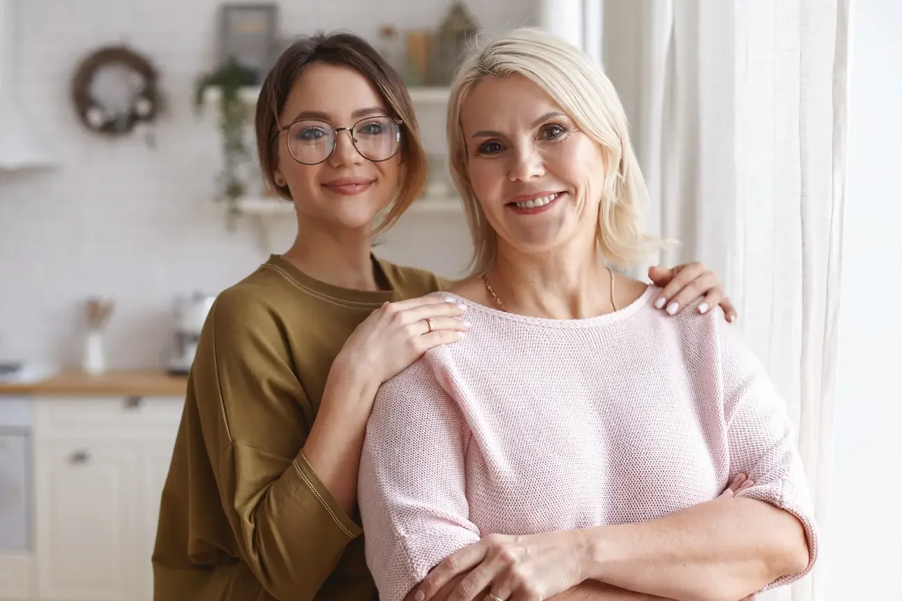 A young woman with glasses and a middle-aged woman with blonde hair smiling, standing close together in a cozy kitchen, with the young woman resting her hand on the older woman's shoulder.
