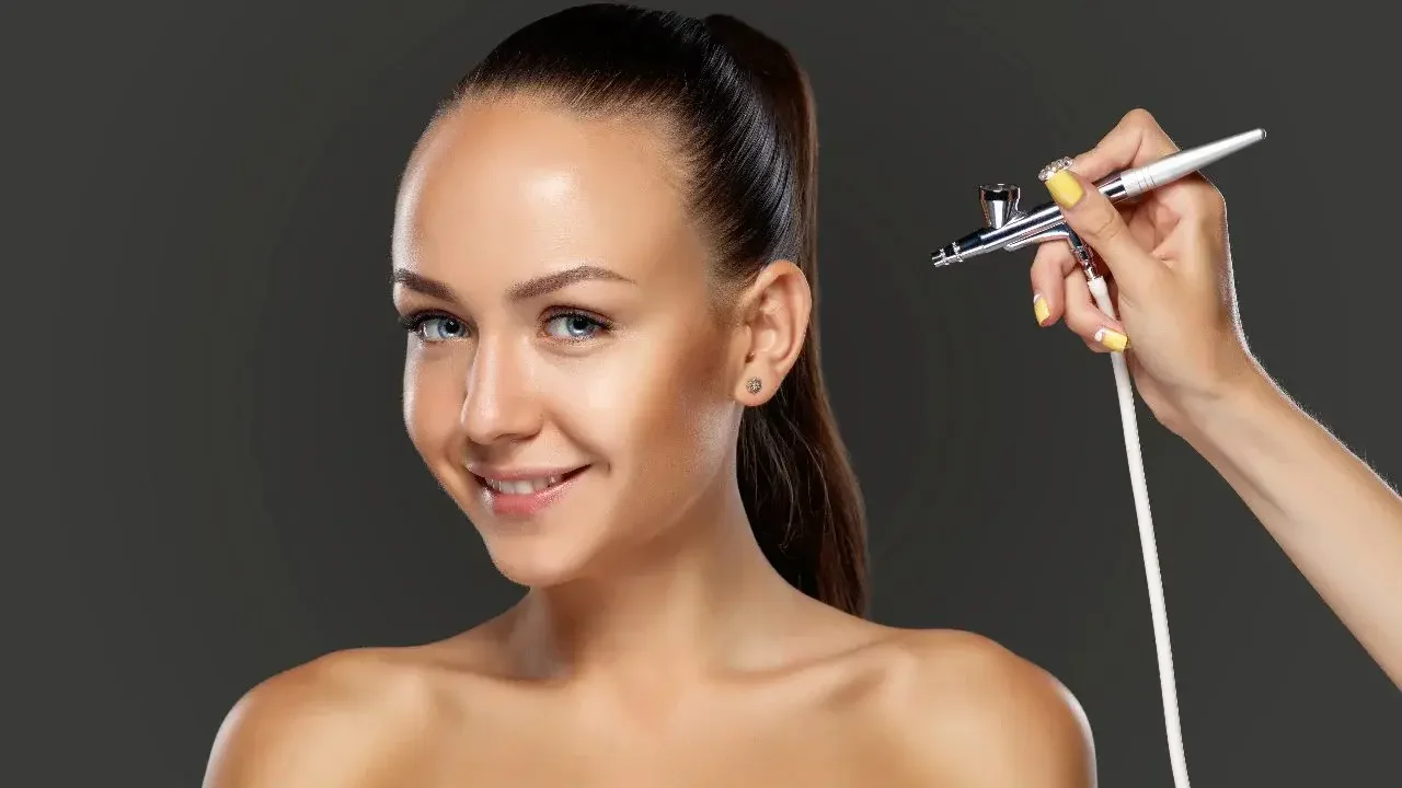 A woman with fair skin and bright blue eyes smiling as an airbrush makeup artist applies makeup on her face. The artist's hand and arm are visible, holding a spray gun connected to a white tube. The woman has her hair pulled back into a ponytail and has small earrings.