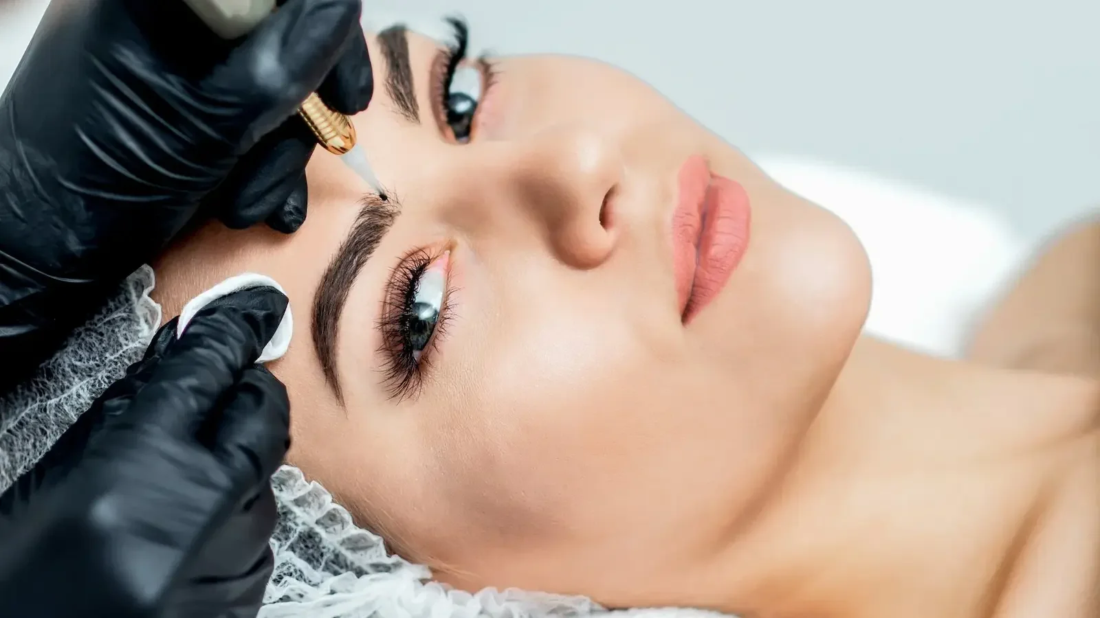 Close-up of a woman receiving a microblading procedure on her eyebrows by a technician wearing black gloves.
