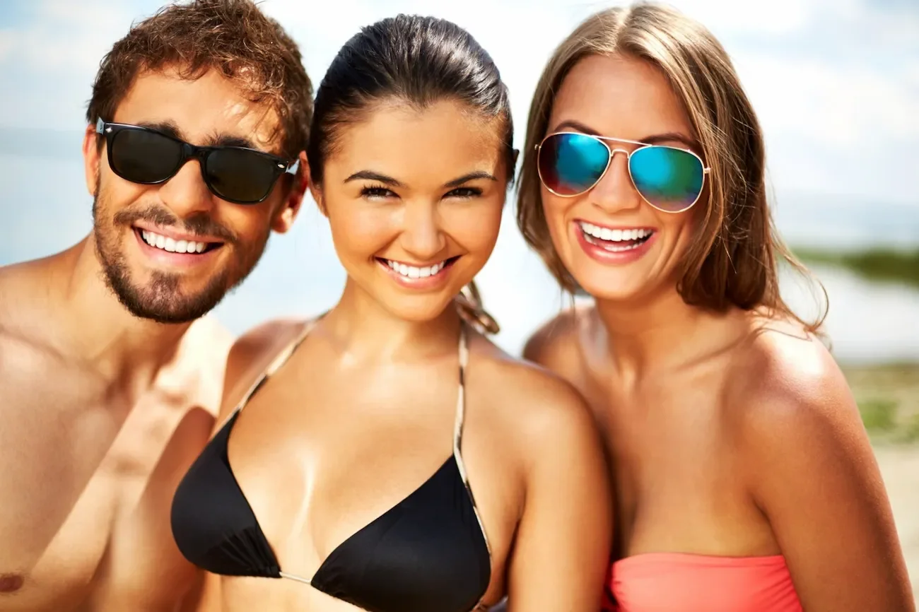 Three young adults smiling and taking a selfie at the beach, wearing sunglasses, in swimsuits, with a blurred ocean and sky background.