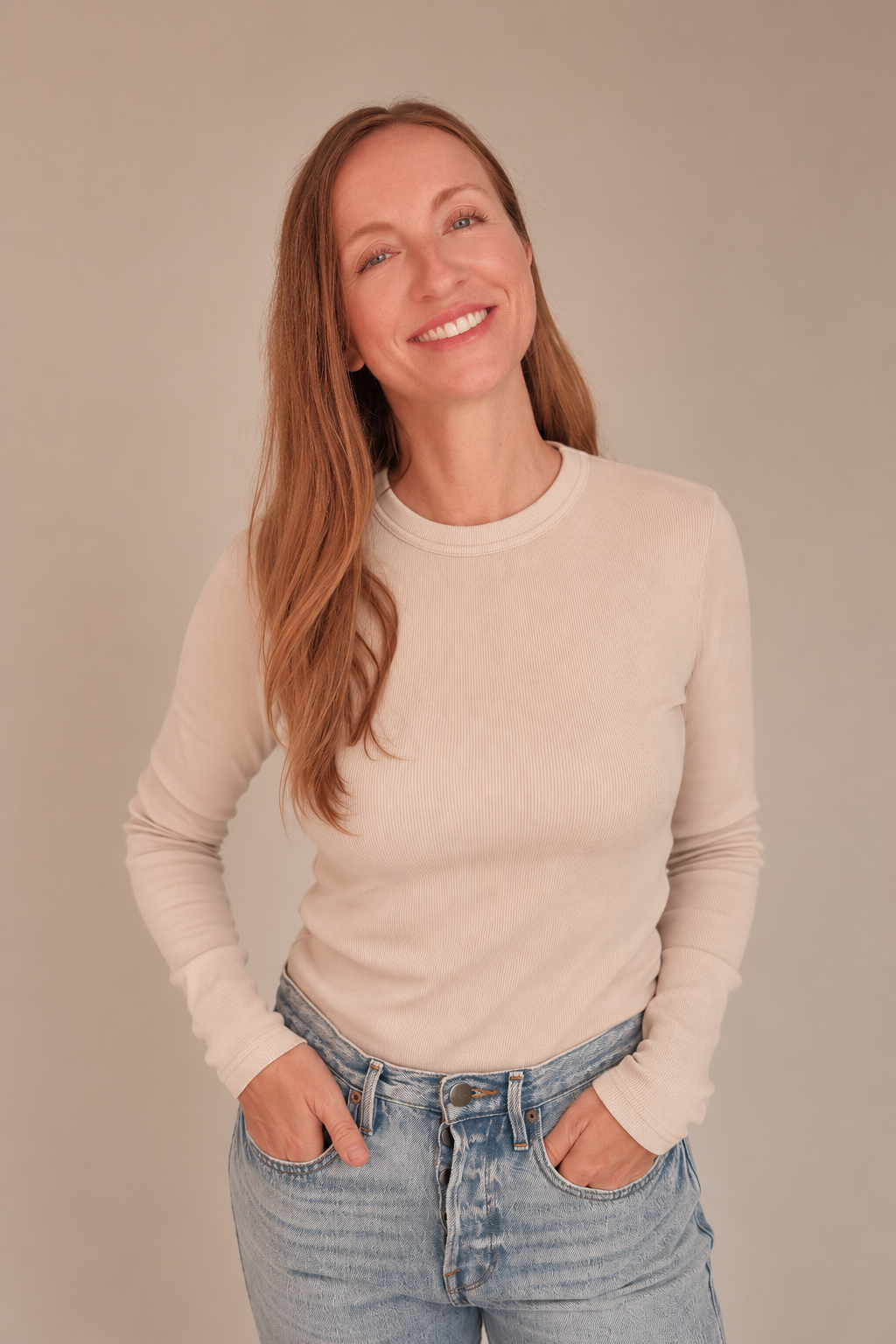 A woman with long red hair, smiling, wearing a beige long-sleeve shirt and light blue jeans, standing against a plain light-colored background.