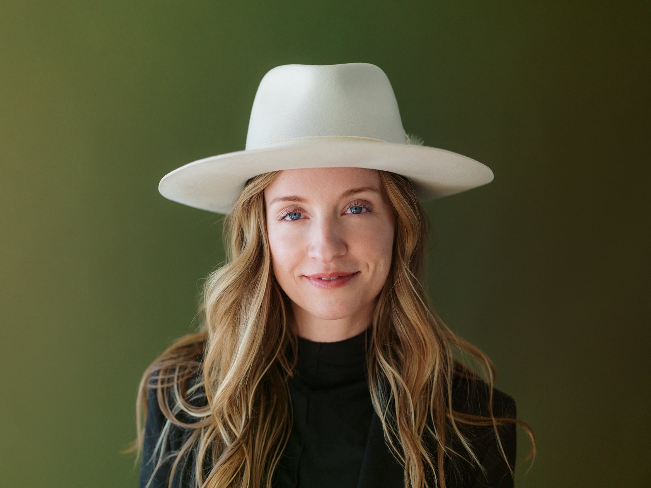 Close-up of a woman with long blond hair wearing a wide-brimmed white hat and a black top with sheer sleeves, smiling softly against a green background.