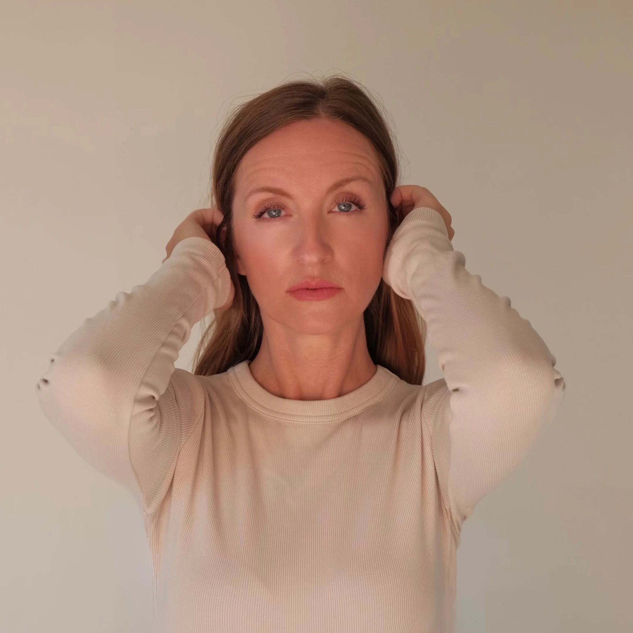 Close-up of a woman with auburn hair, light blue eyes, wearing a beige ribbed sweater, and placing her hands behind her ears, against a plain neutral background.