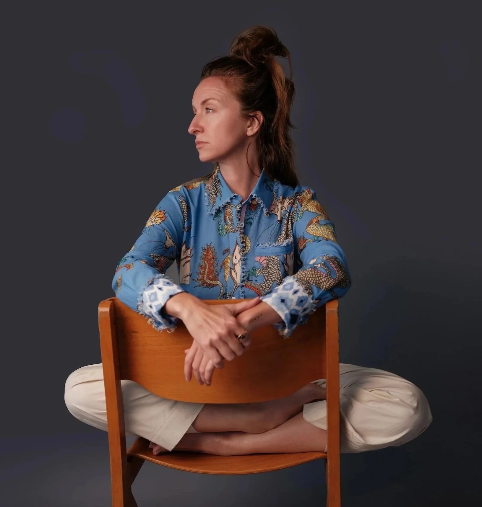 A woman with brown hair tied in a messy bun, sitting cross-legged behind a wooden chair, wearing a blue patterned shirt and beige pants, against a dark gray background.