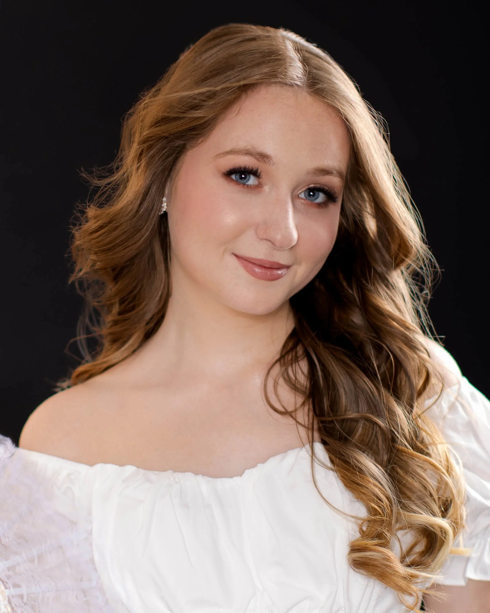 A woman with long, wavy, light brown hair, blue eyes, and light skin, wearing a white off-shoulder top and earrings, smiling softly against a black background.