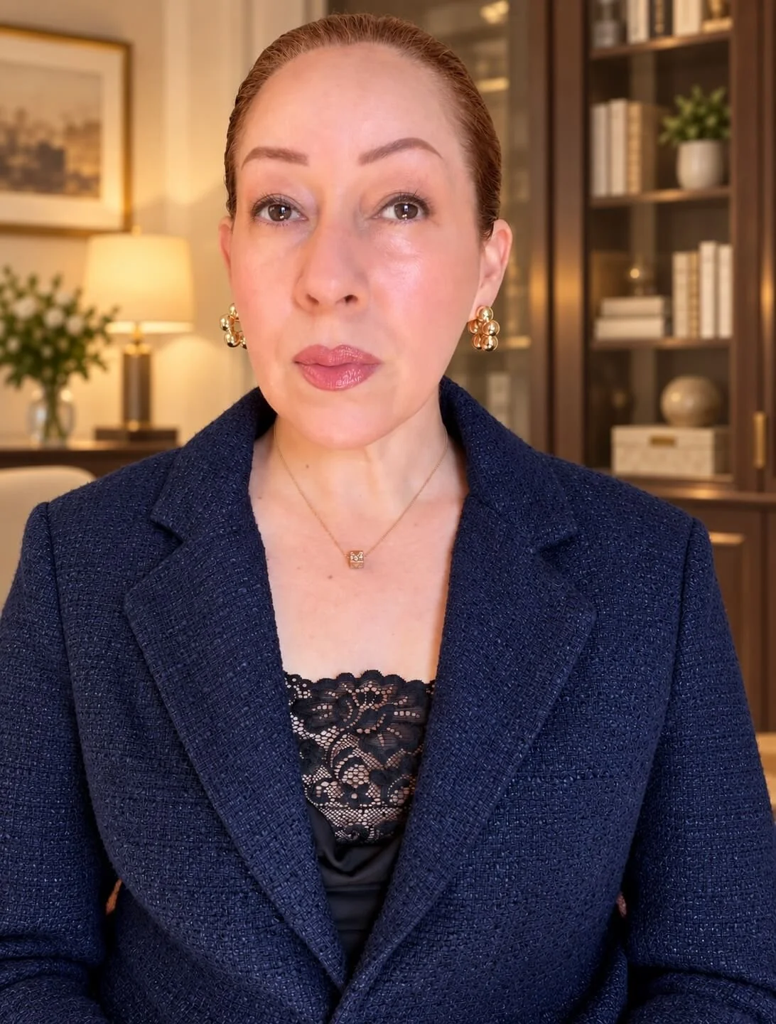 Close-up of a woman with fair skin, red hair pulled back, wearing a navy blazer, gold earrings, and a necklace, in a warmly-lit room with bookshelves and a lamp in the background.