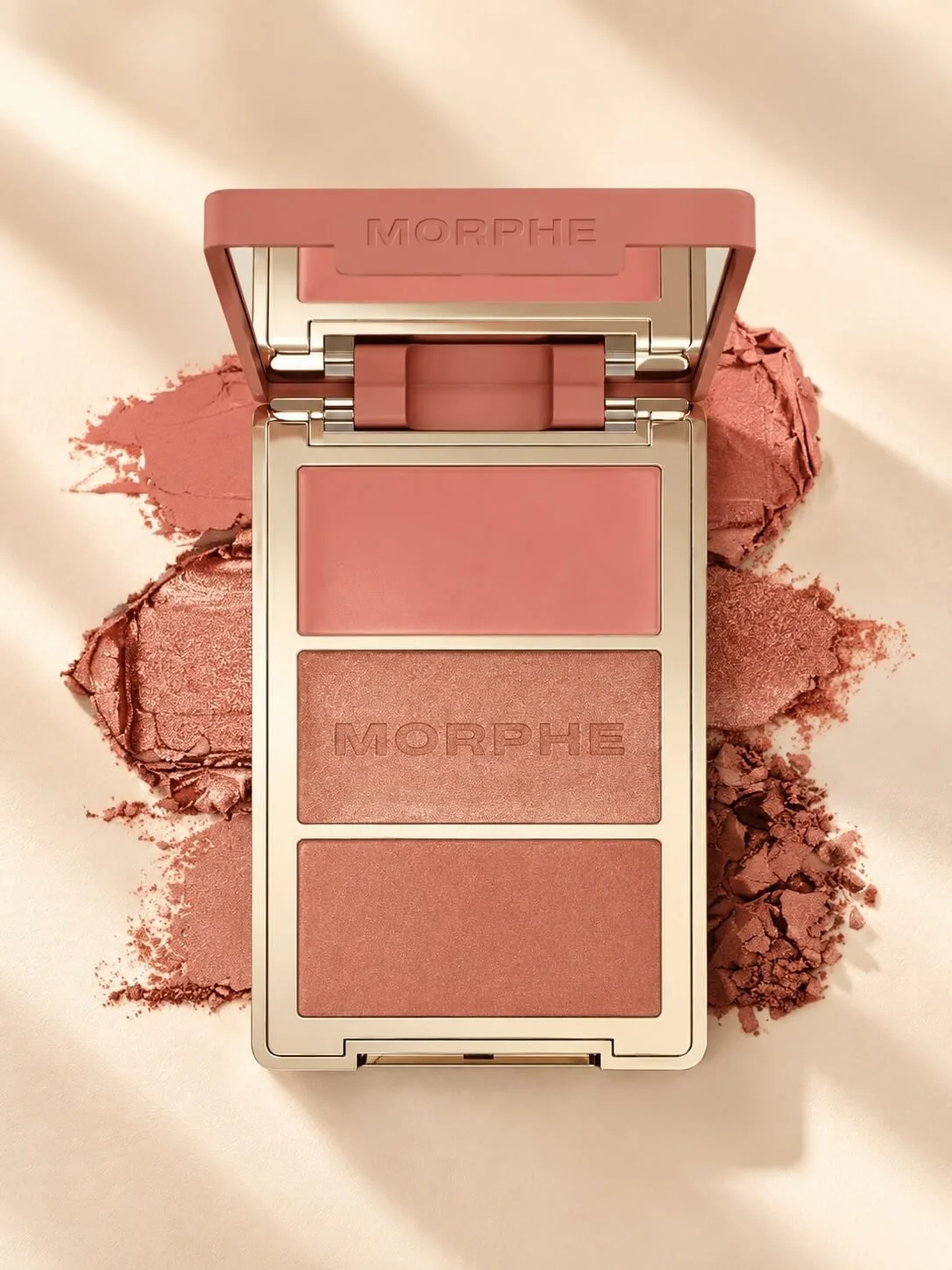 A makeup palette with three different shades of pink and bronze pressed powders, branded with 'MORPHE,' on a soft pink surface with spilled powder around it.
