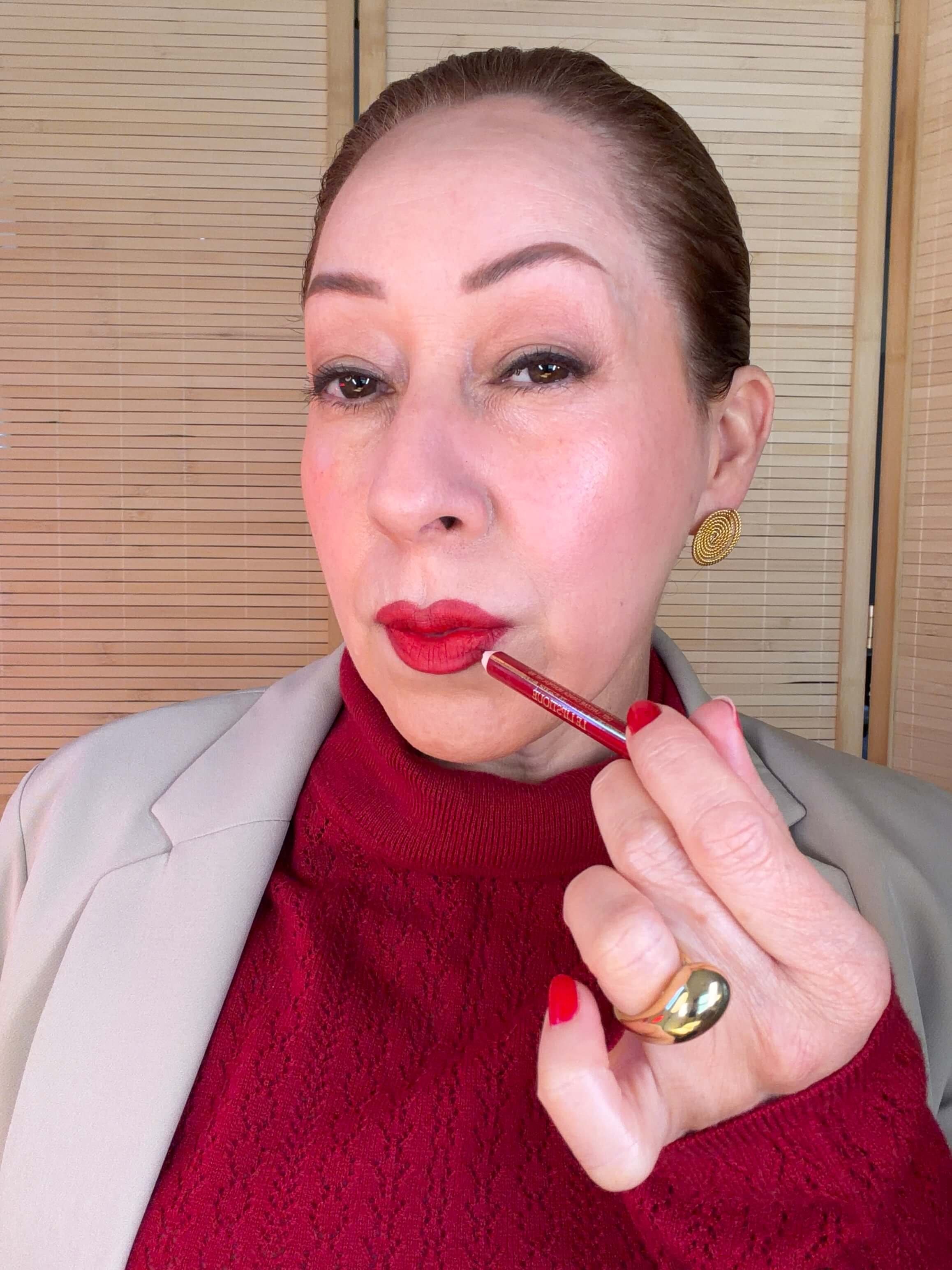 María with red lipstick and gold earrings applies red lipstick with a pencil. She is wearing a red knit top and a beige blazer, with a background of wooden panels.