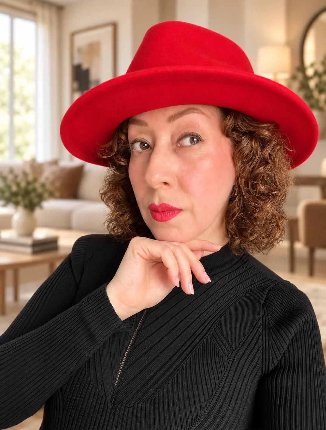 María wearing a red wide-brimmed hat and red lipstick, posing indoors with her hand under her chin.