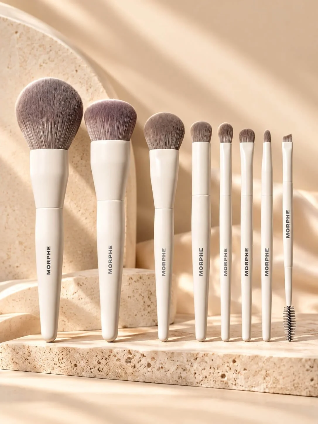 A collection of Morphe makeup brushes with white handles and light-colored bristles, displayed on a beige stone surface.