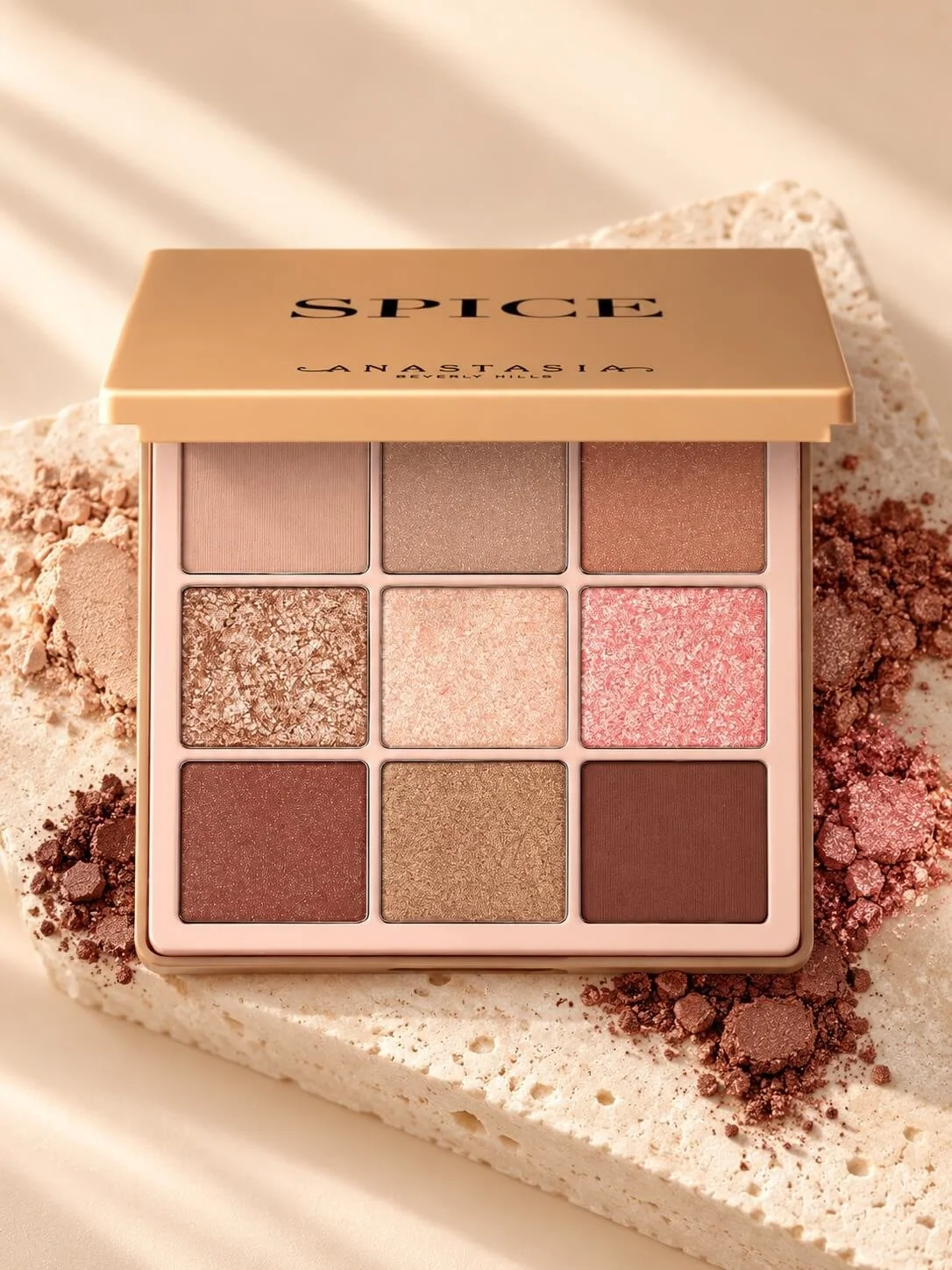 An Anastasia Beverly Hills Spice eyeshadow palette with nine shades in neutral and pink tones, resting on pink and brown powdered crumbs on a light surface.
