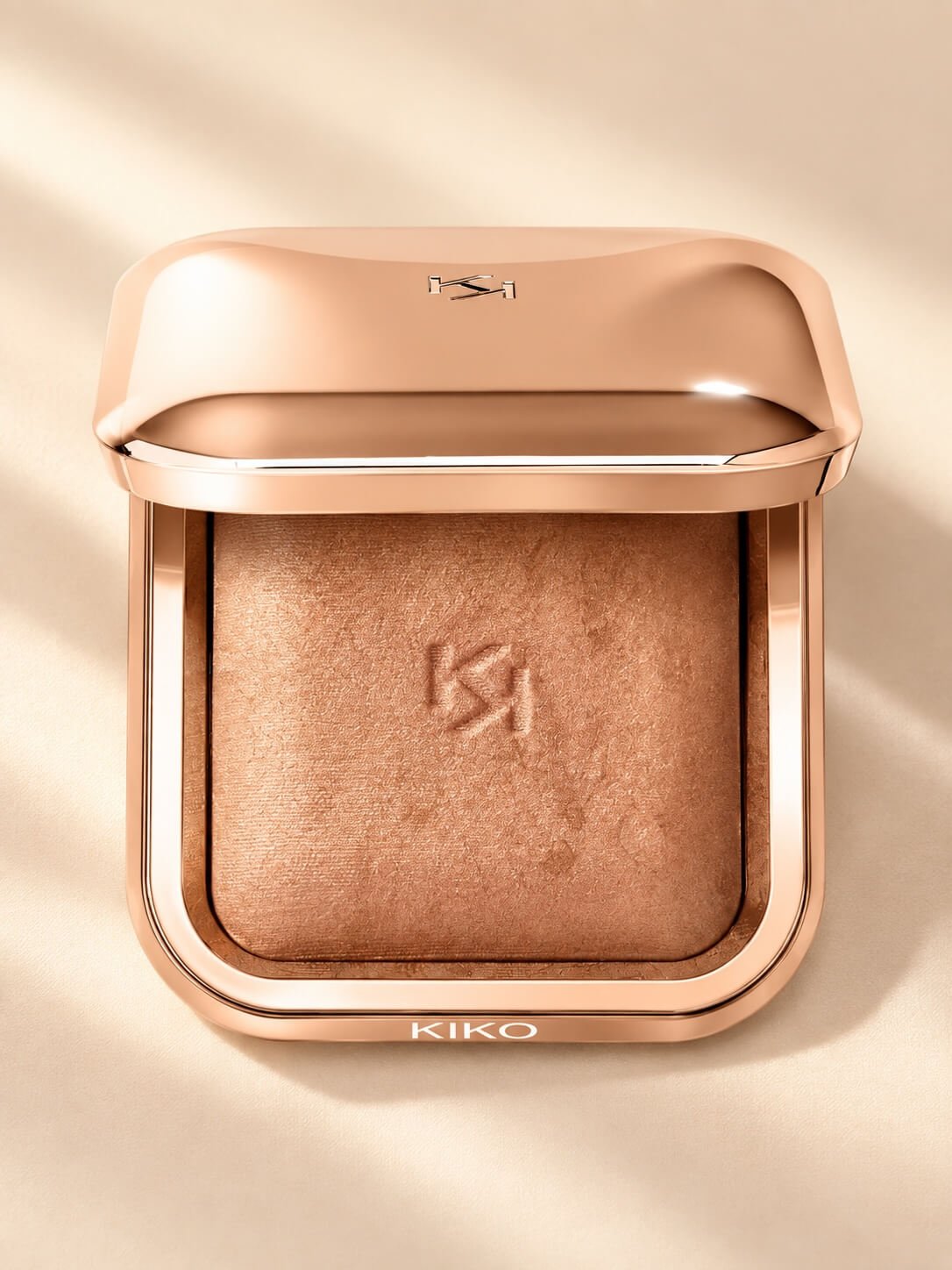 A jar of KIKO makeup with a metallic peach-colored lid and a shimmery peach eyeshadow inside, embossed with the KIKO logo, set against a smooth, light-colored background.