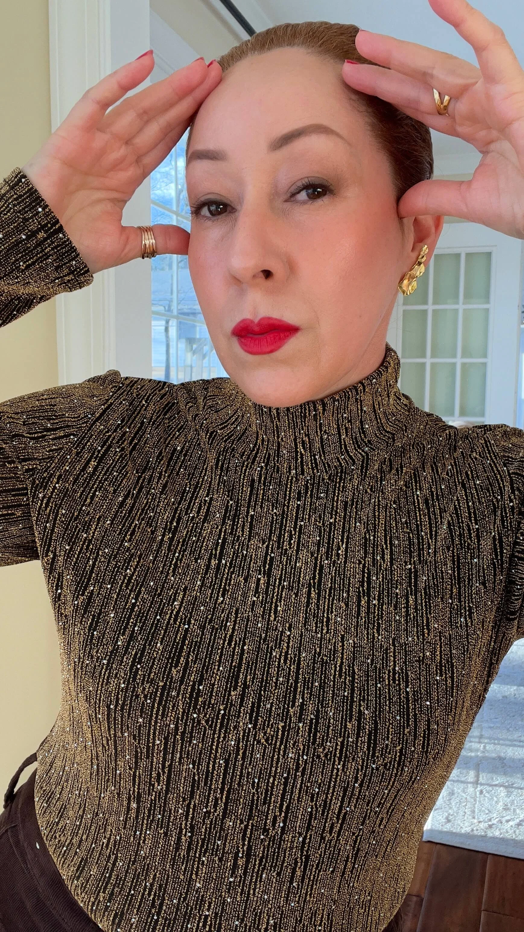 María with red hair styled back is wearing a gold and black glittery turtleneck top, large gold earrings, and rings. She is standing indoors near a window and door, posing with her hands near her head.