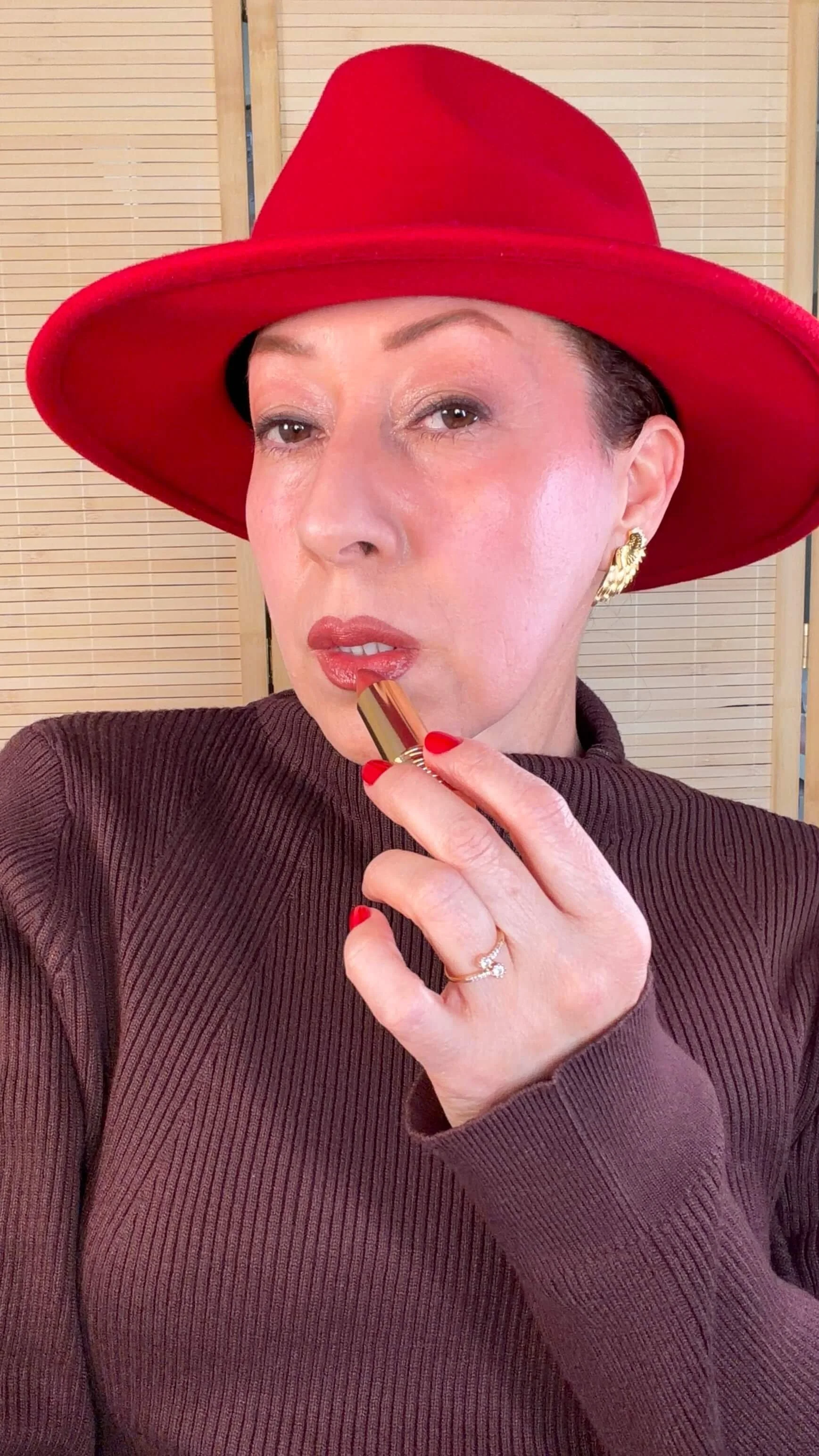 María with red hat applying lipstick, wearing a brown ribbed sweater and gold earrings, with a wooden panel background.