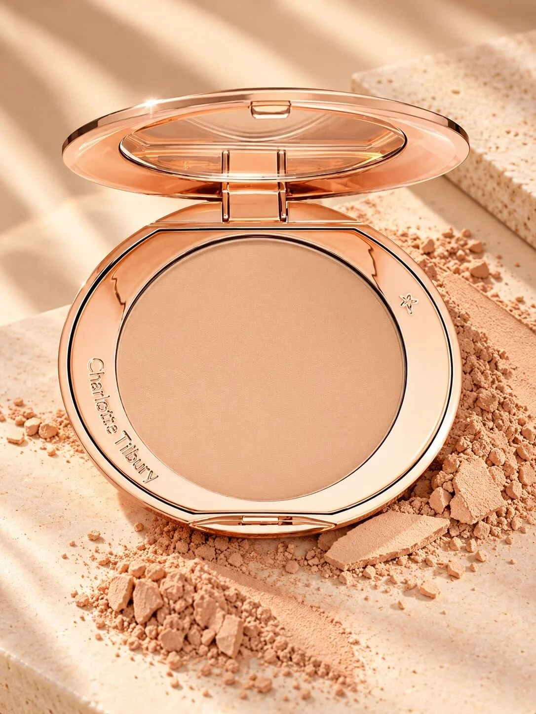 A compact pressed powder makeup case with a rose gold finish, open to reveal a beige powder, surrounded by loose powder and powder fragments on a beige textured surface.