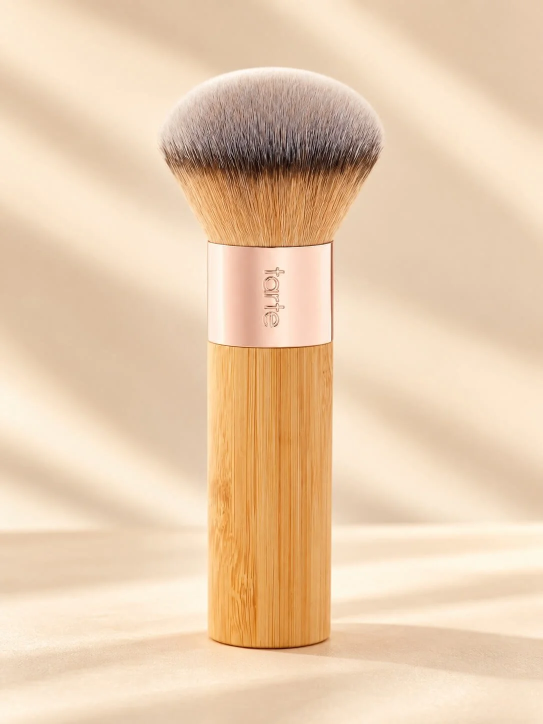 A makeup brush with a wooden handle, a rose gold ring engraved with the word 'tarte,' and soft, fluffy bristles against a beige background.