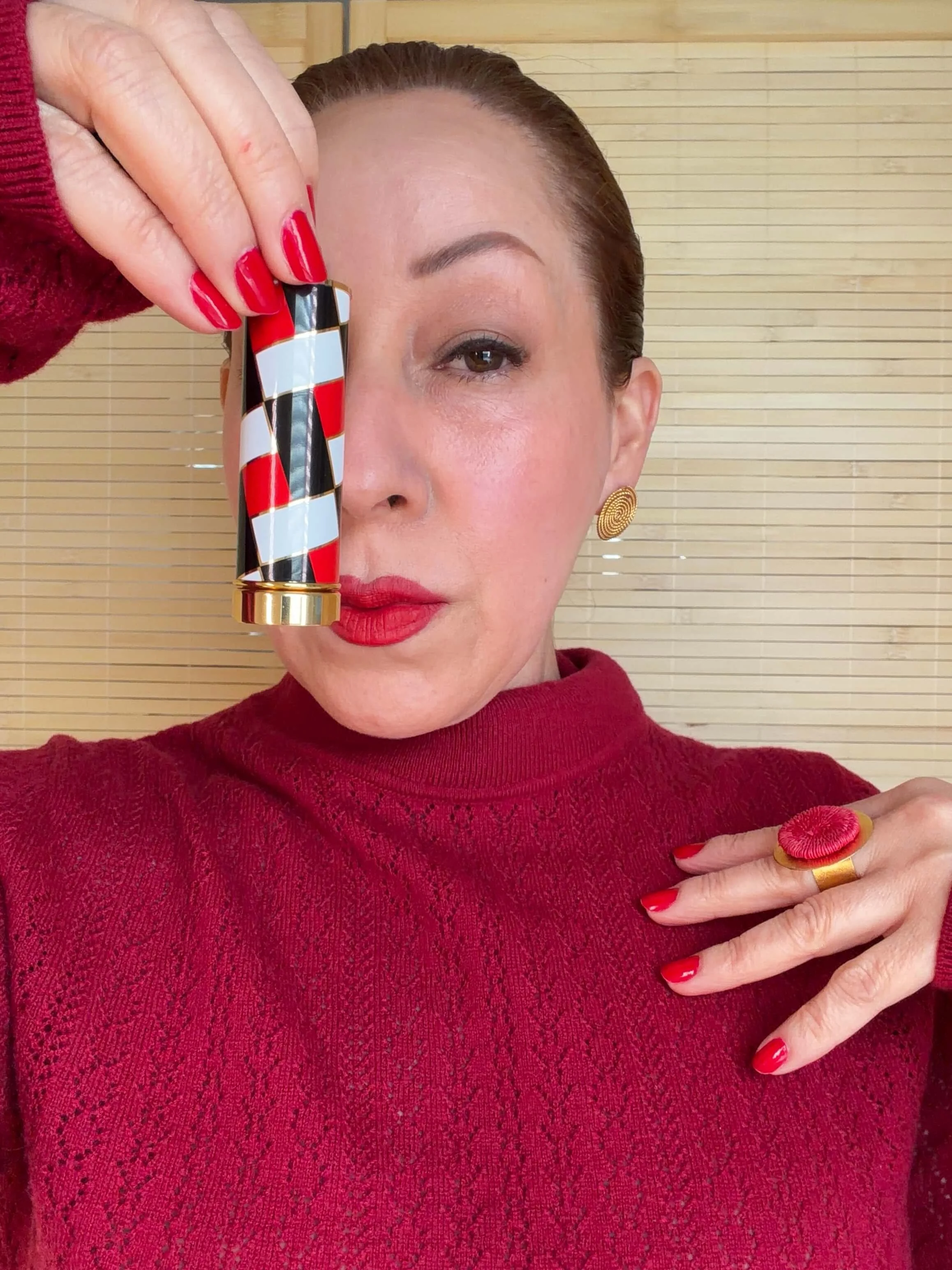 María with red lipstick and red painted nails holds a patterned lipstick tube near her face, wearing gold earrings, a red sweater, and a large red and gold ring. She is standing in front of a wooden background.