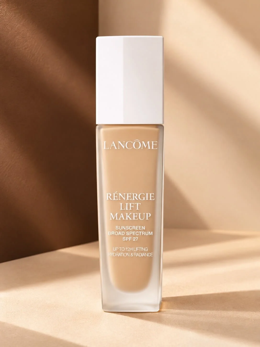 A bottle of Lancôme Rénergie Lift Makeup with SPF 27, positioned on a light-colored surface against a backdrop of beige and brown tones.