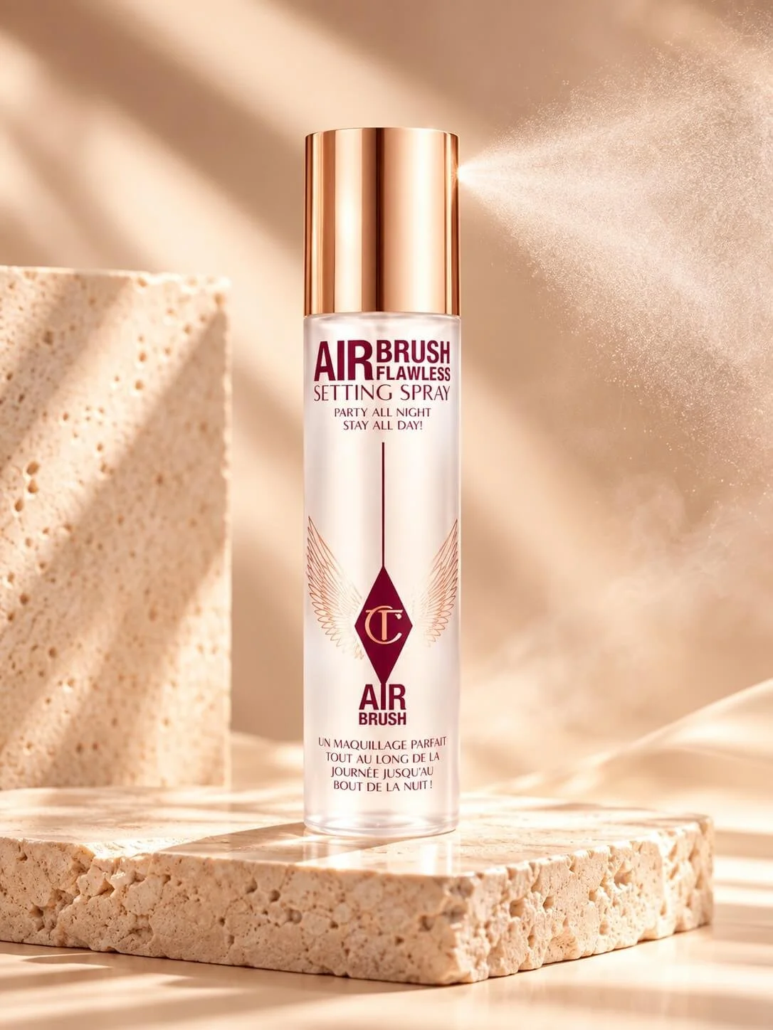 A bottle of Air Brush Flawless Setting Spray with a rose gold cap, placed on a textured stone surface with a backdrop of soft beige fabric and shadows.