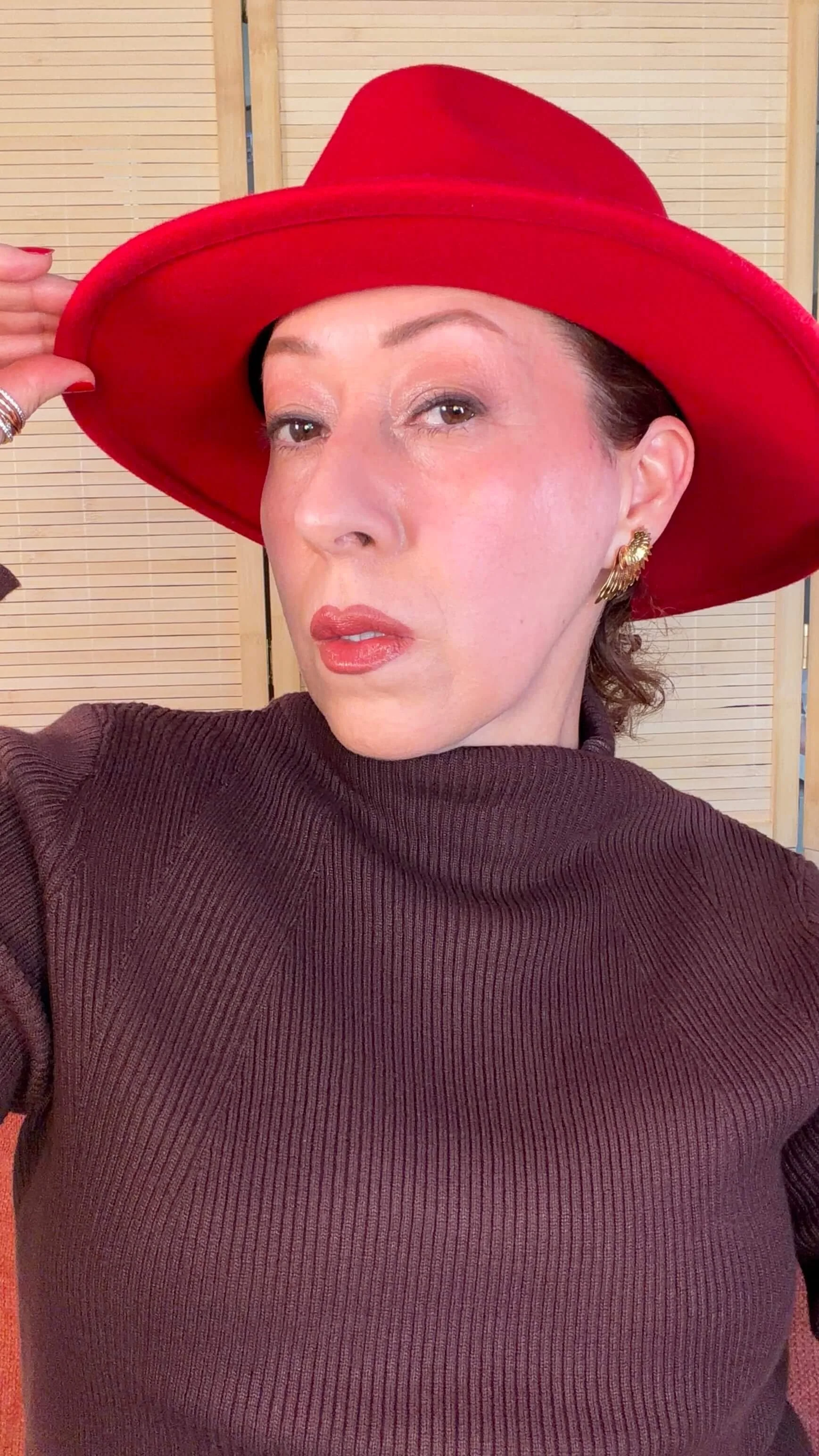 María taking a selfie wearing a red hat, gold earrings, and a dark brown ribbed turtleneck sweater, with a light-colored background.