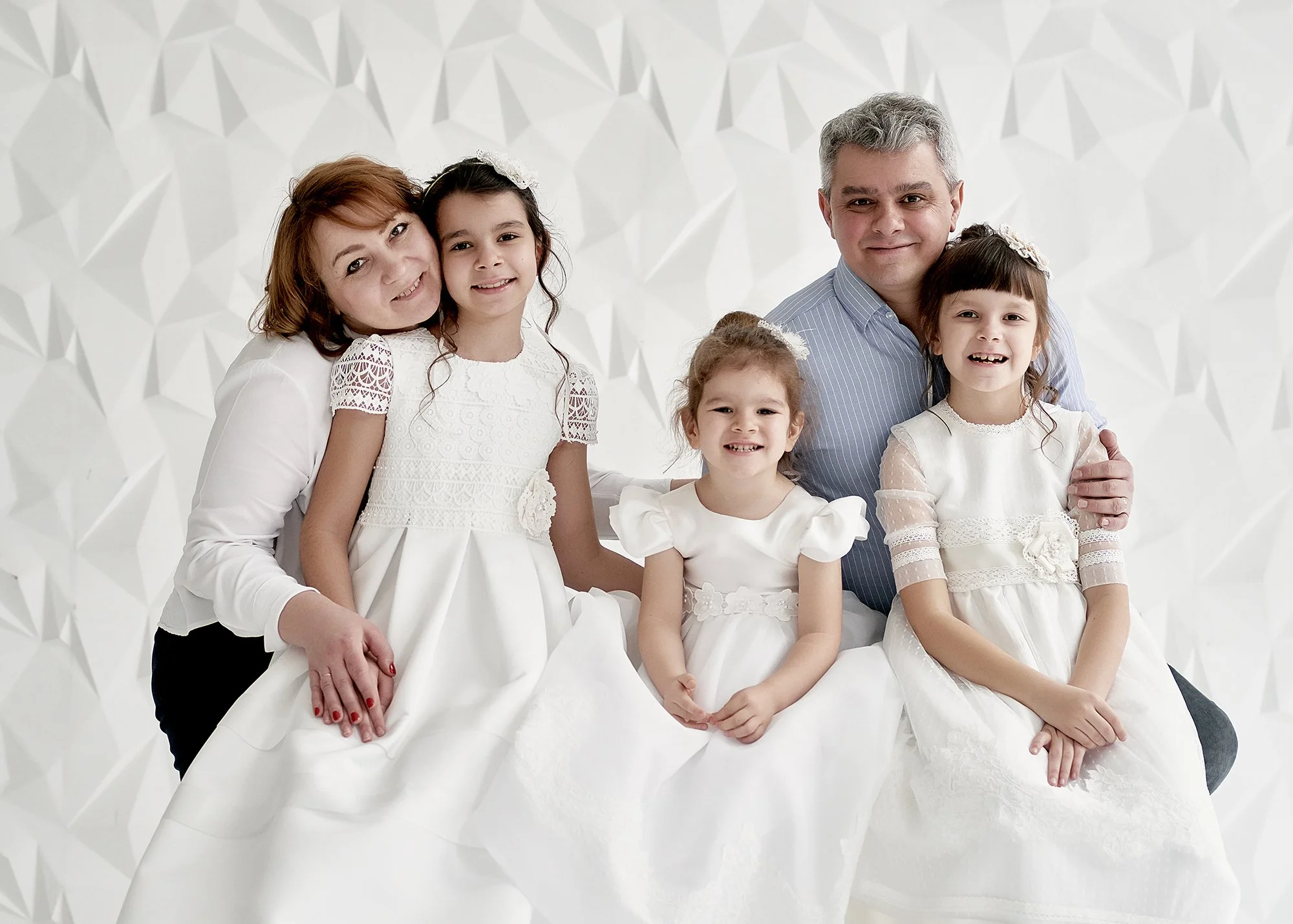 Family portrait with a man, woman, and three children in white dresses against a geometric white background.