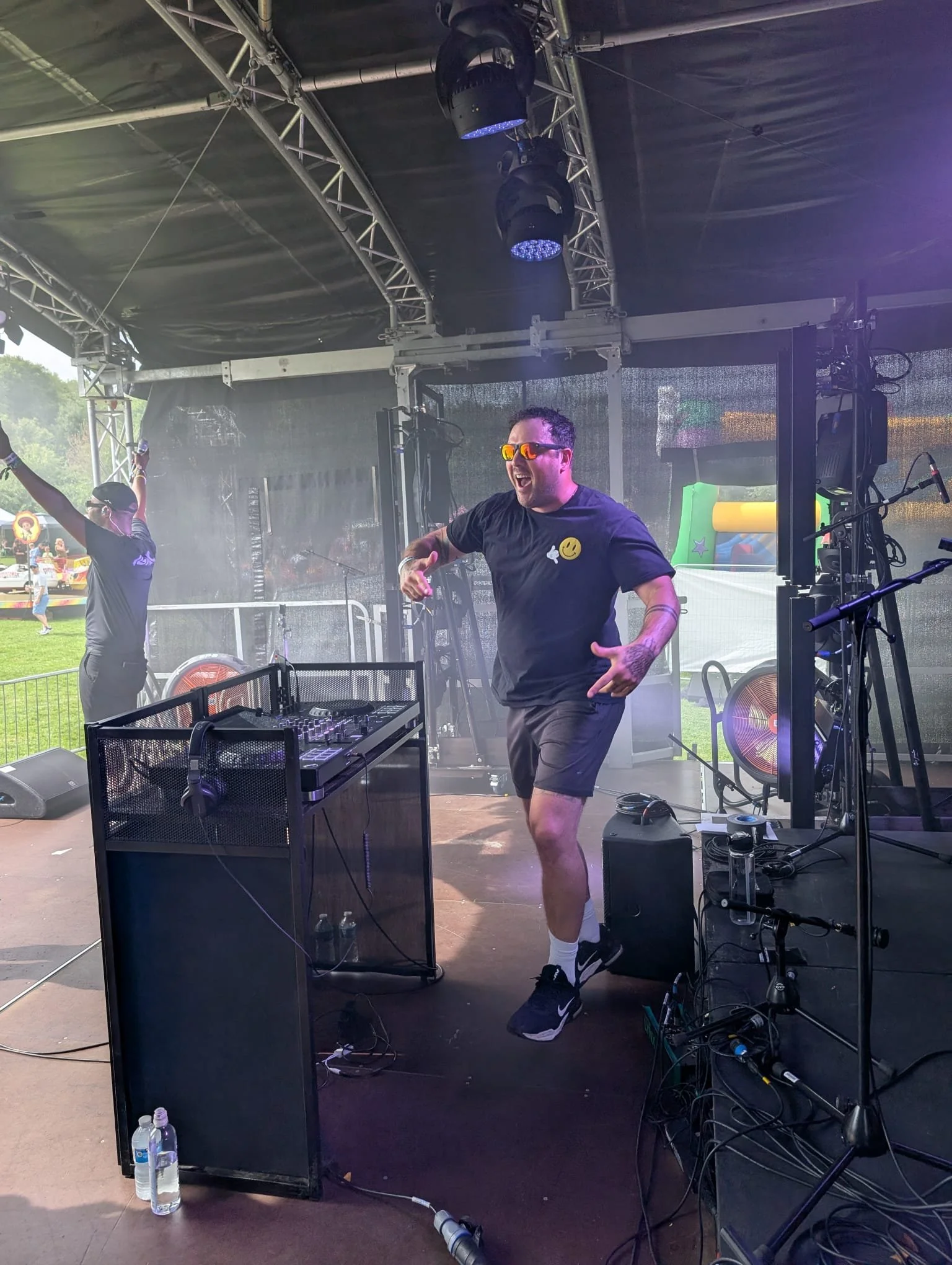 A DJ performs at an outdoor event under a large tent, wearing sunglasses and a black shirt with smiley and star patches, with colorful lighting, sound equipment, and people in the background.