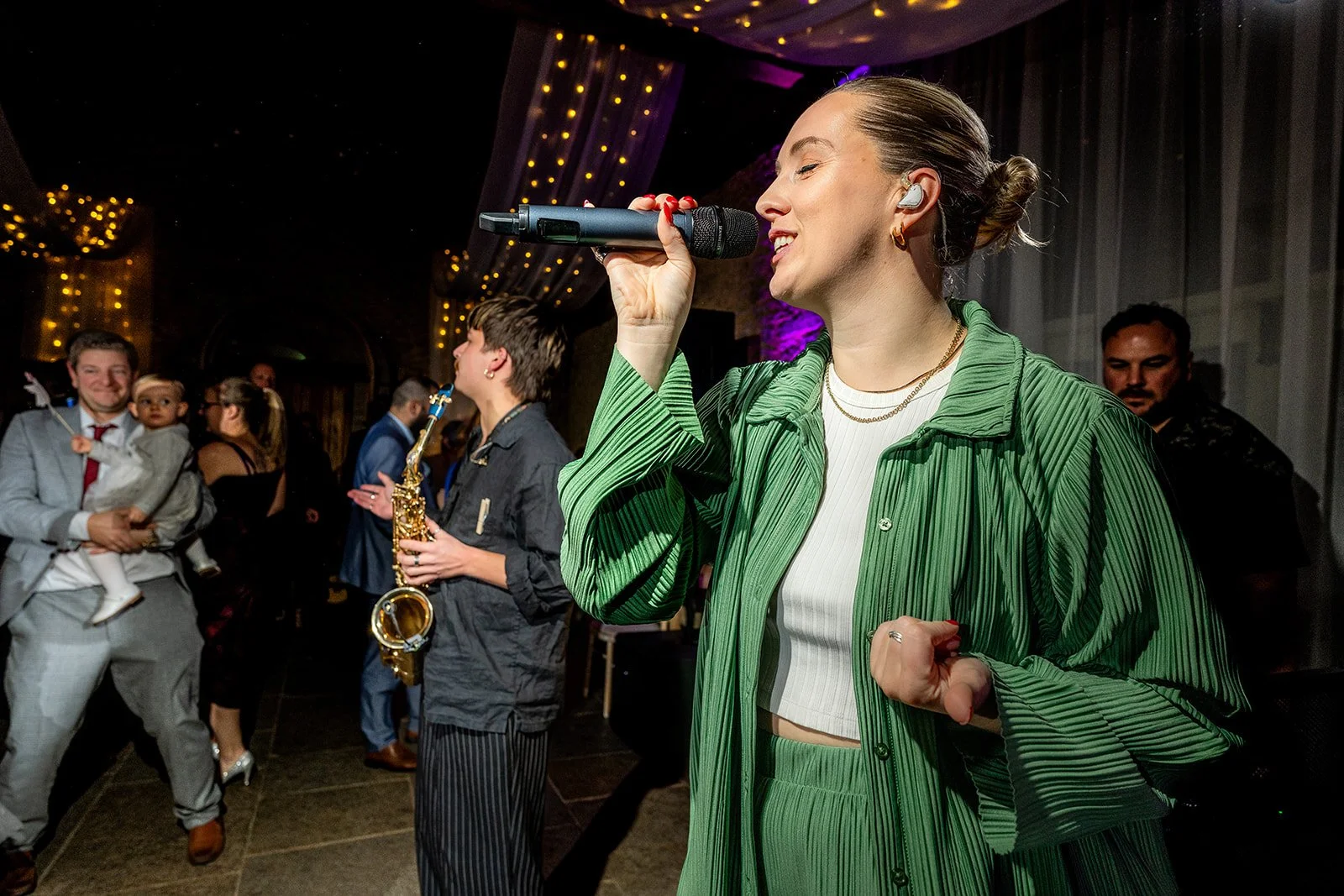 A woman with styled hair singing into a microphone at a party or event, with a crowd and some musicians, including a young man holding a saxophone, in the background.