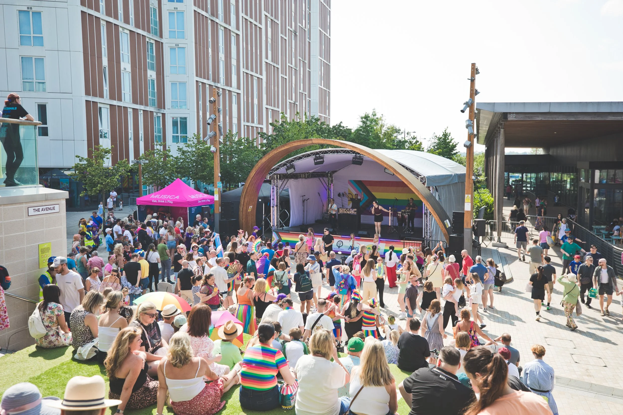 A large outdoor crowd gathered in front of a stage with a rainbow-colored backdrop, with many sitting on the grass or standing, at a festival or event during sunny weather in an urban area with modern buildings nearby.