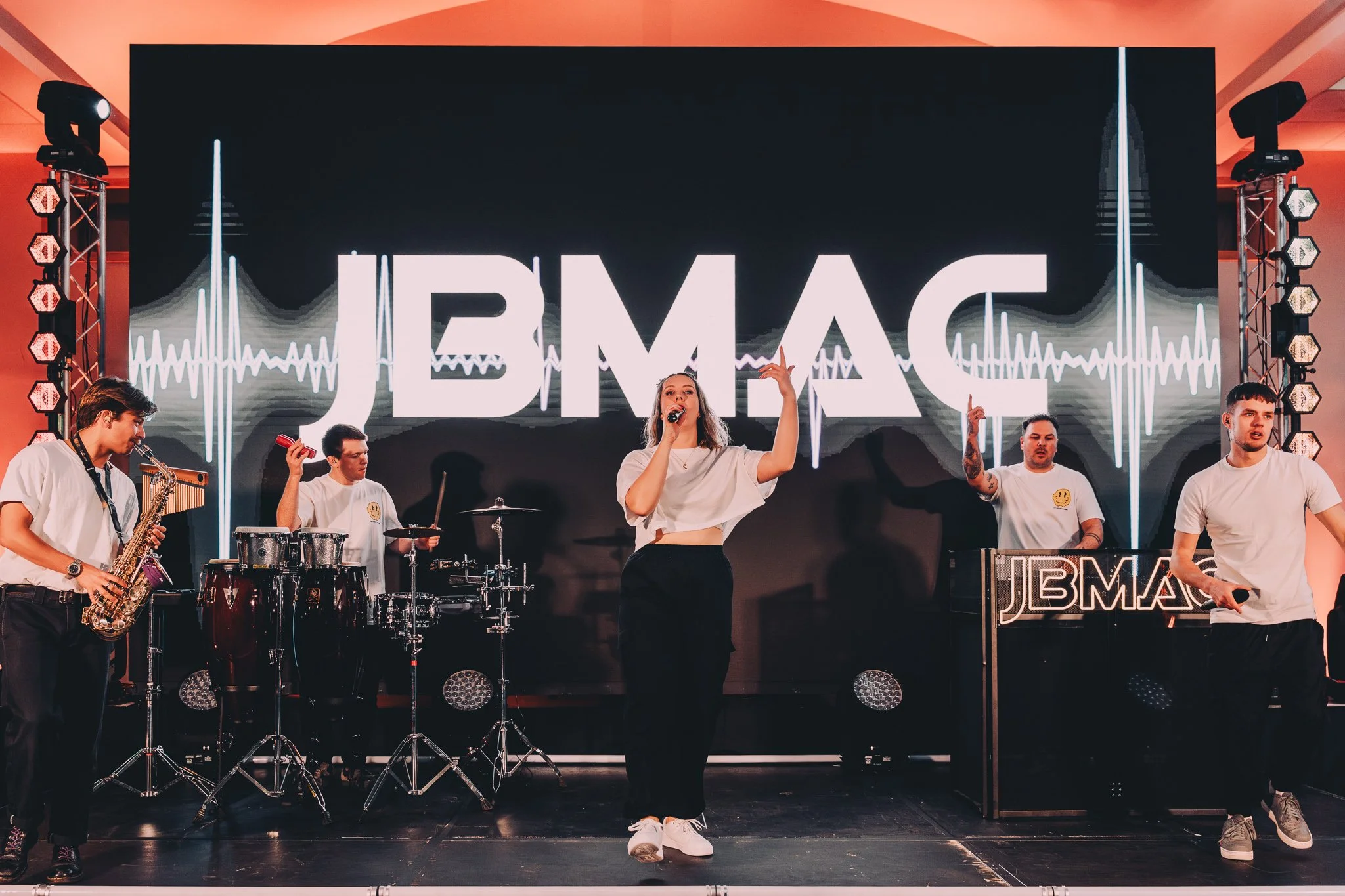 A live music performance with five band members on stage, and a large LED screen behind them displaying the word 'JBMAC' with soundwave graphics.