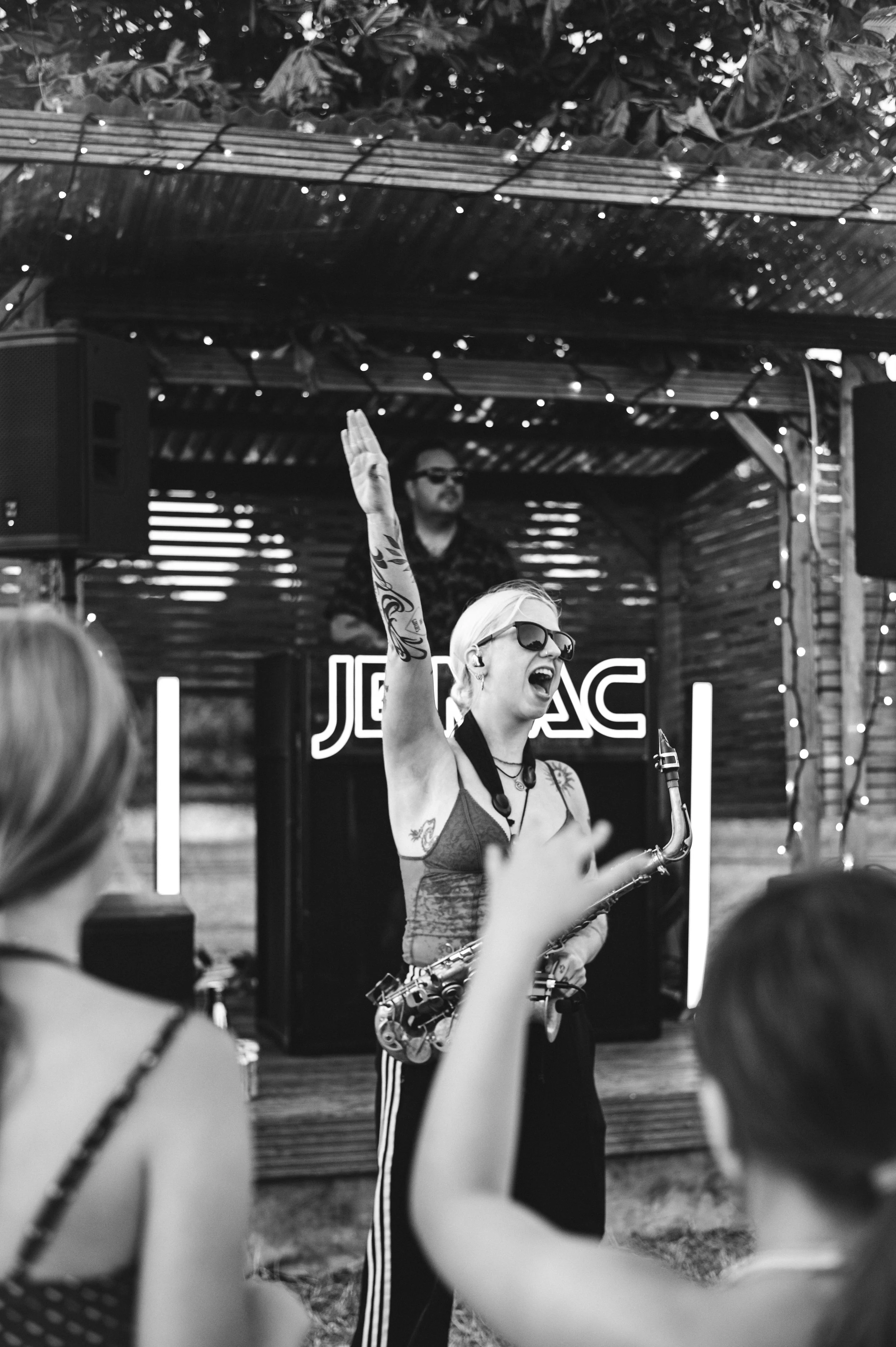 A woman with tattoos, sunglasses, and short blond hair playing a saxophone, raising her hand in front of a small audience outdoors. A DJ behind her in the background, with a sign that says 'JACK' in neon lights.