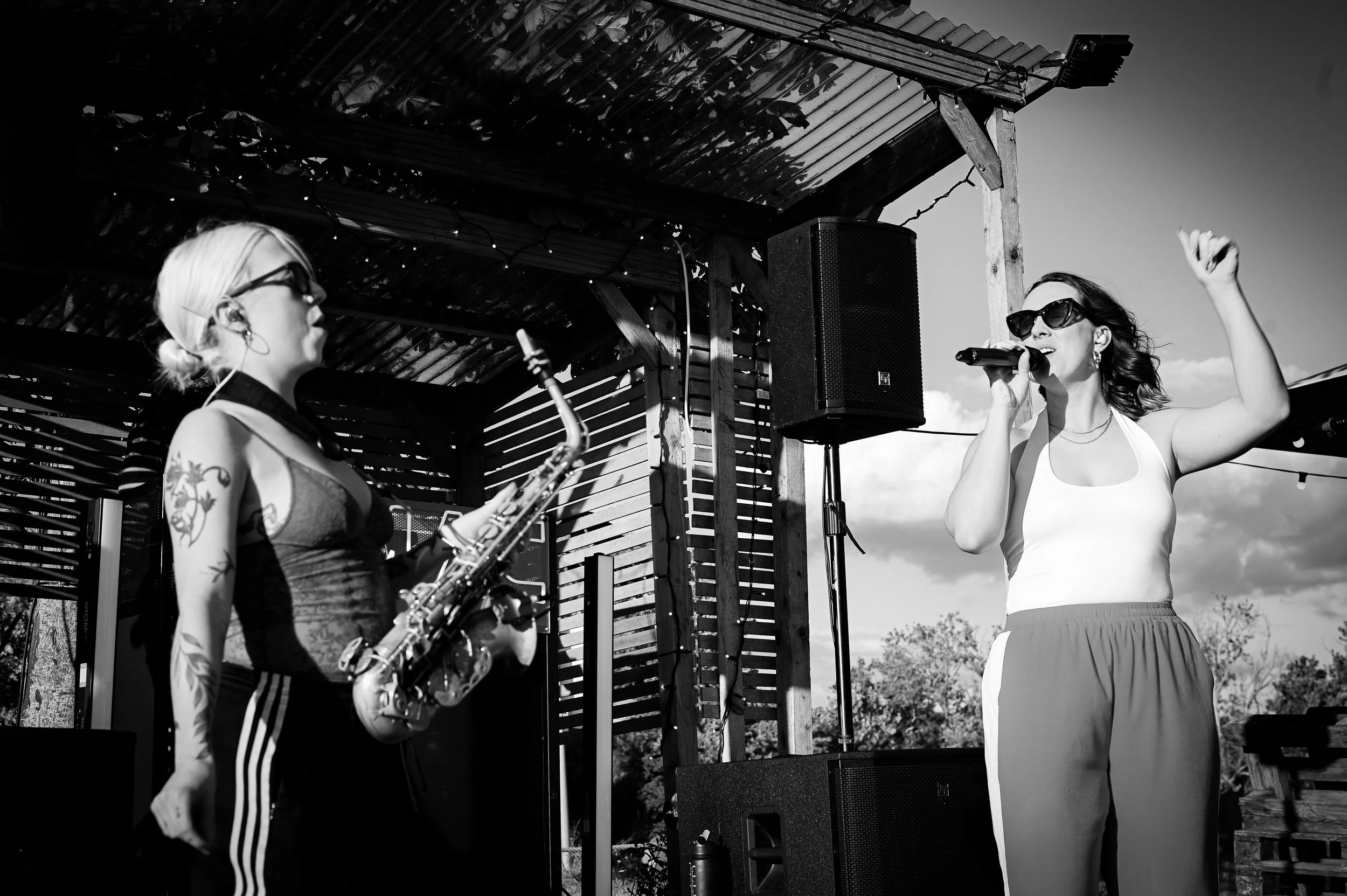 Two women performing on an outdoor stage, one singing into a microphone and the other playing a saxophone, under a wooden shelter with string lights, in black and white.