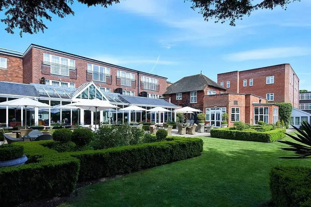 A well-maintained outdoor garden with neatly trimmed hedges and lawn, multiple patio tables with chairs, white umbrellas, and a large building with glass conservatories and brick architecture under a bright blue sky.