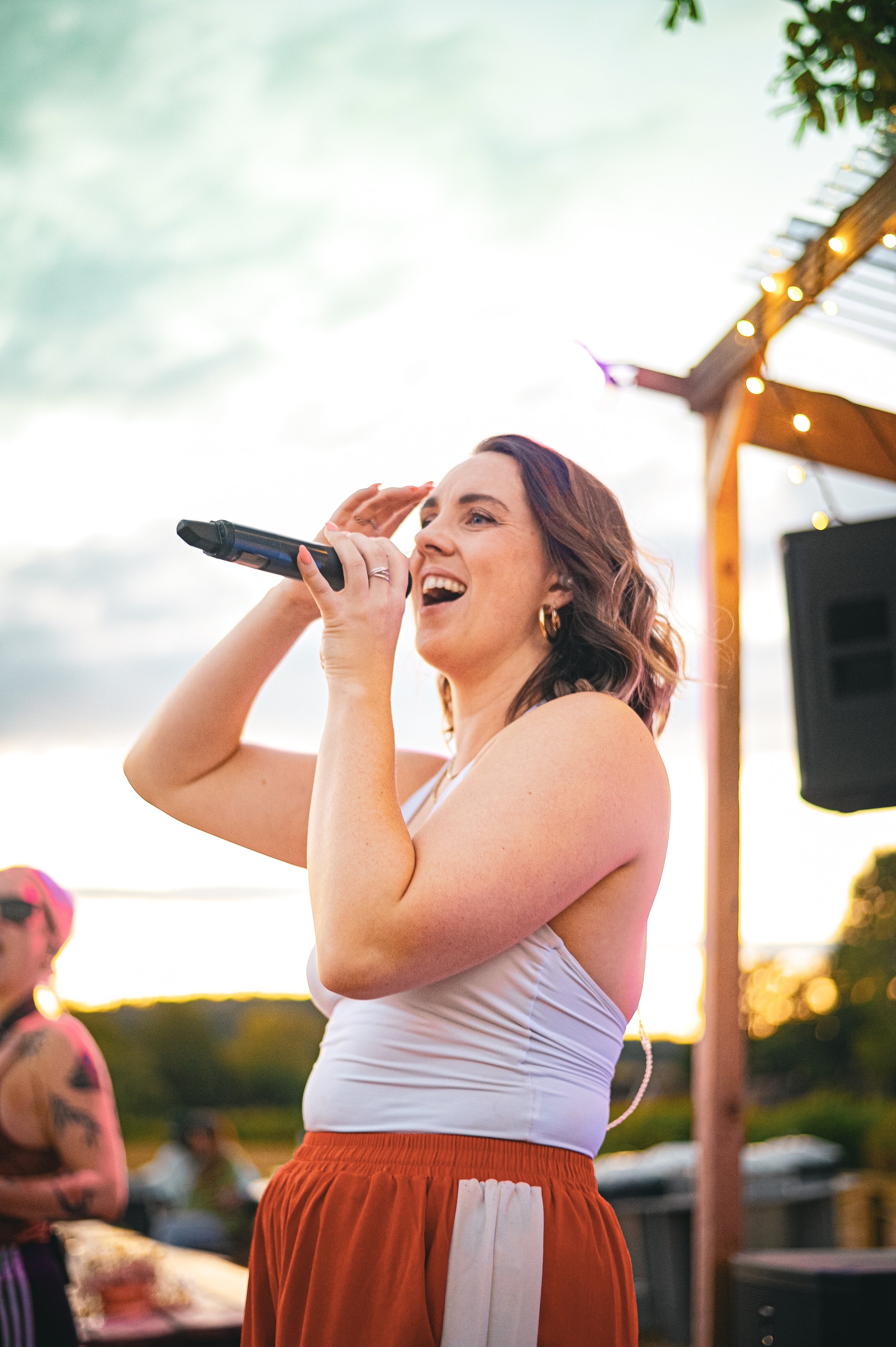 A woman with wavy brown hair singing into a microphone at an outdoor event during sunset. She is wearing a white strapless top and orange shorts. There are trees and a wooden structure with string lights in the background.