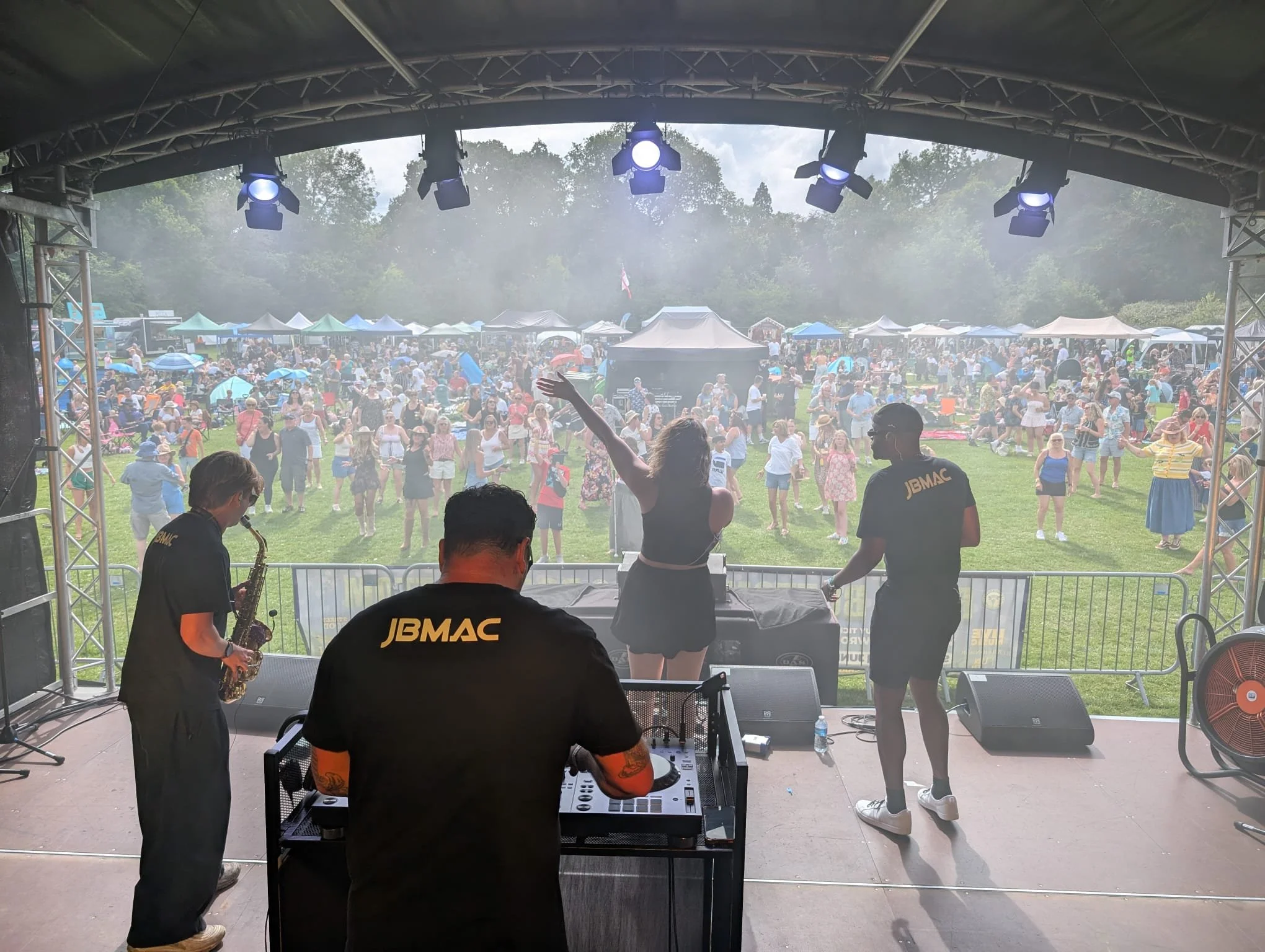 Musicians perform on a stage at an outdoor event with a large crowd under tents and umbrellas in a park. The photo is taken from behind the band.