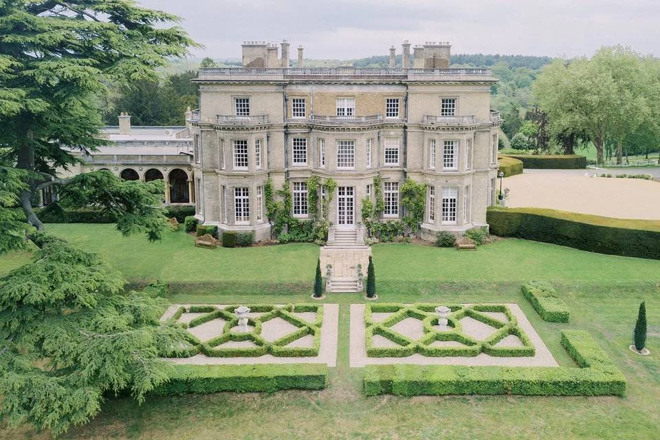 Large historic mansion with a symmetrical garden front, trimmed hedges, and a lawn surrounded by trees and open land.