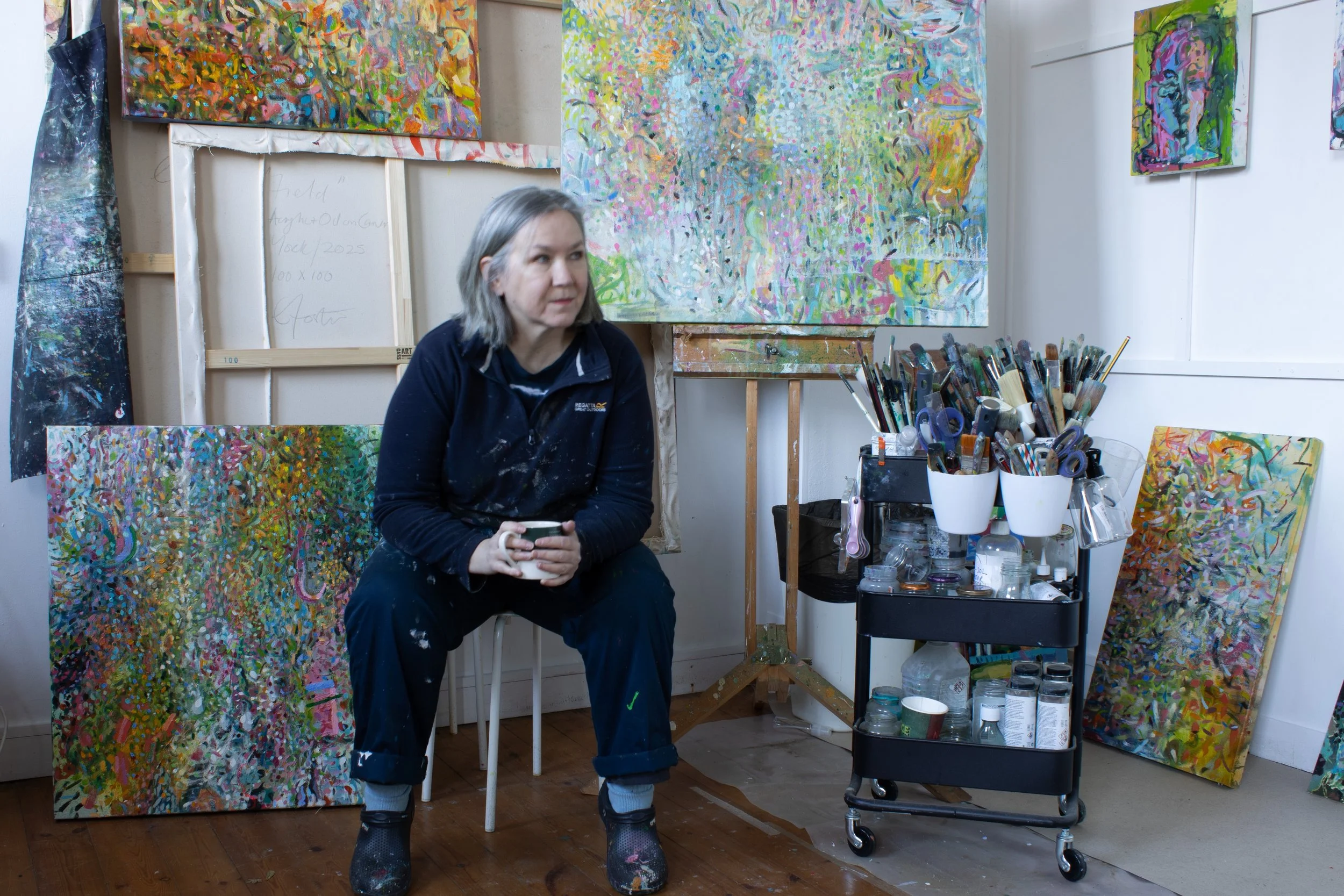 A photograph of the artist Liz Foster siting in her studio with a mug of coffee in her hand looking towards some work that is out of shot.