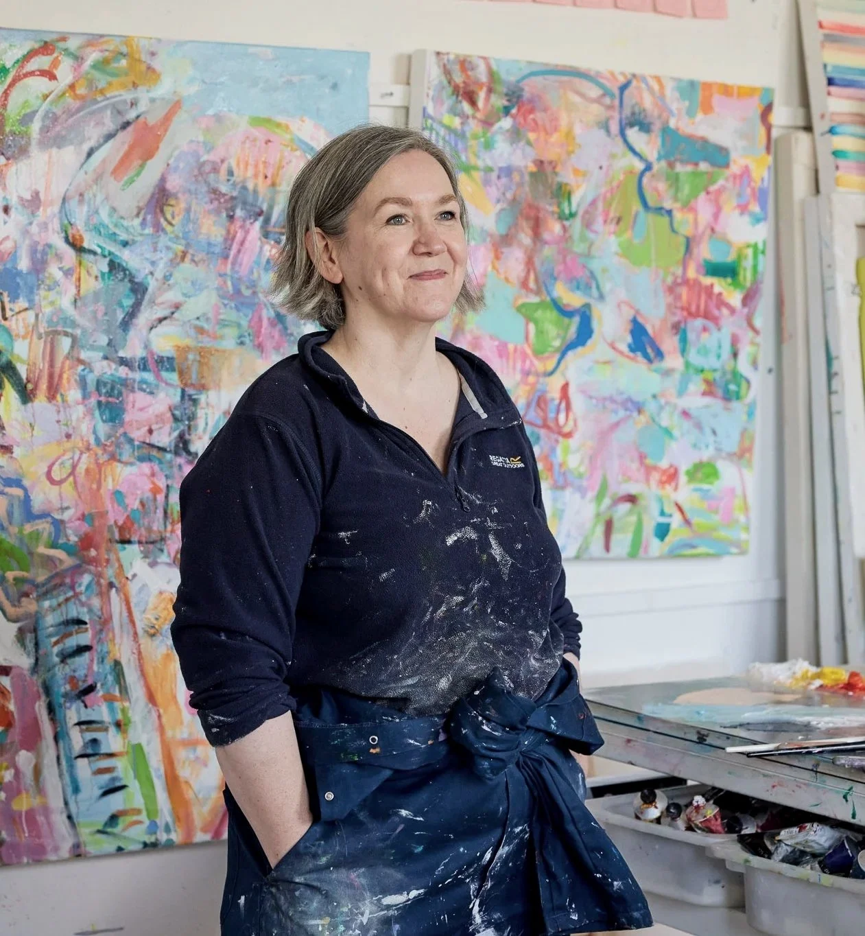 A photograph of the artist LIZ FOSTER standing in her studio, hands pockets with wet paintings hanging on the wall behind her.